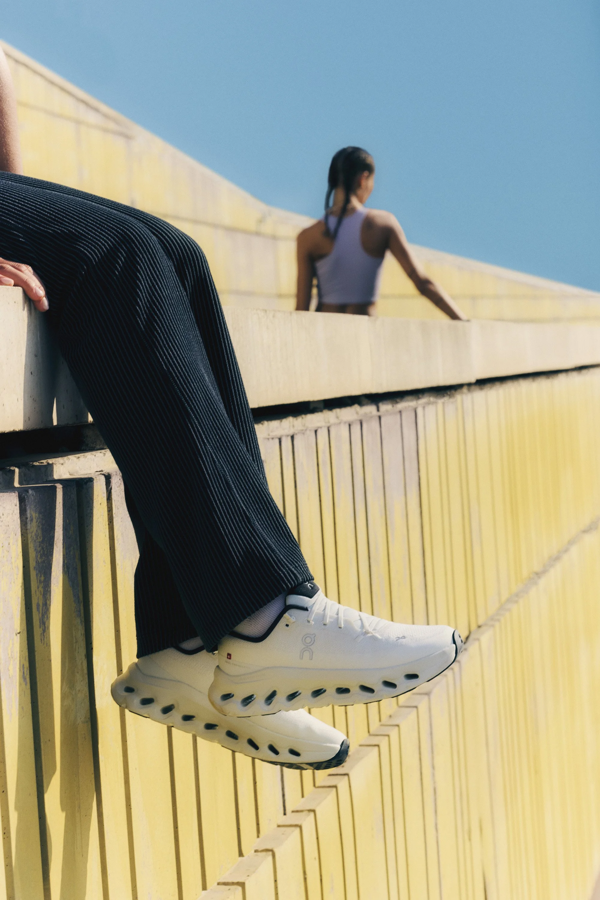 Close-up of a person's legs wearing black pinstripe pants and white athletic shoes, hanging over a yellow railing, with a woman in a white tank top in the background against a clear blue sky.