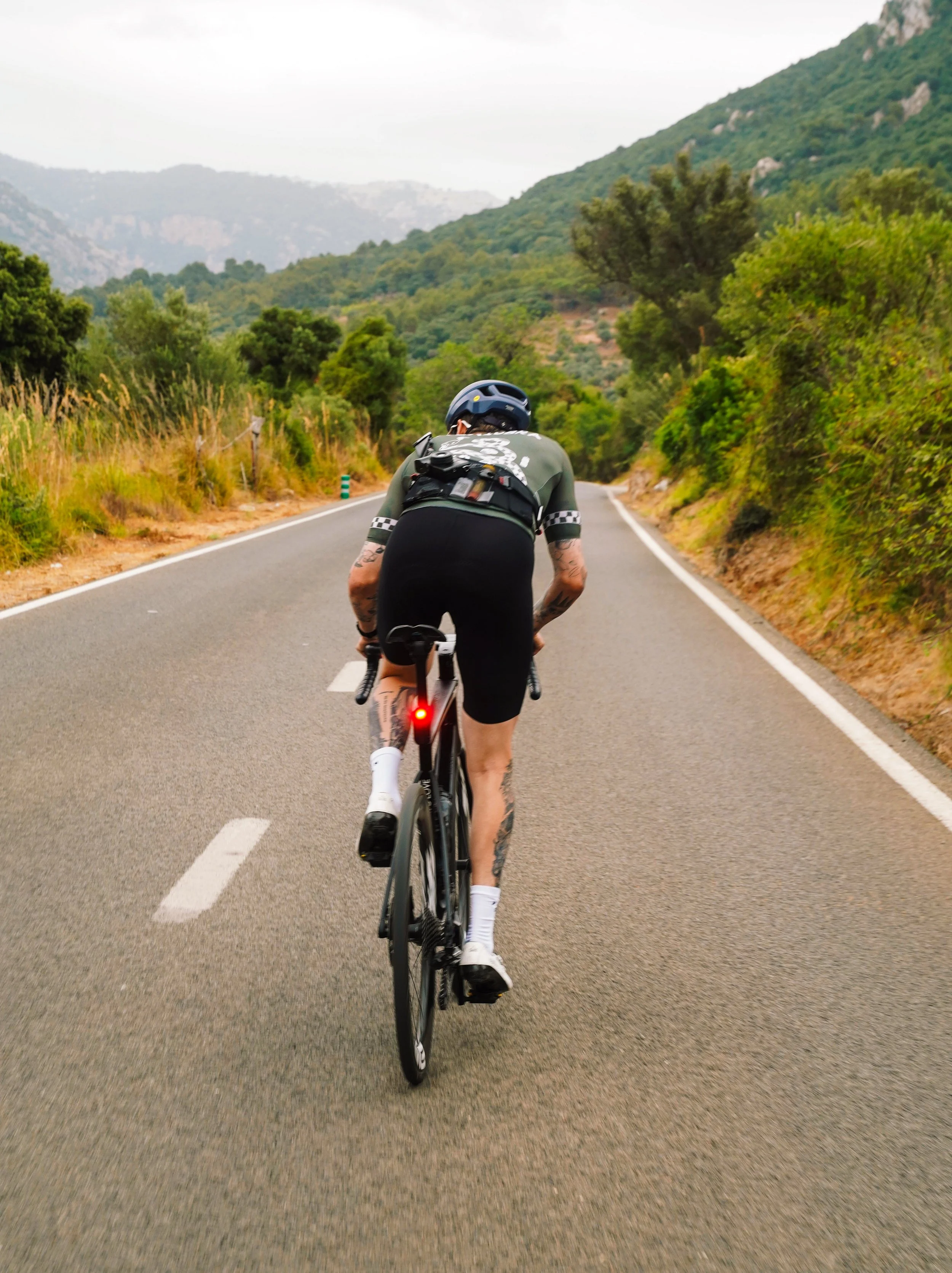 A cyclist wearing a helmet and black shorts riding on a paved mountain road surrounded by green trees and hills.