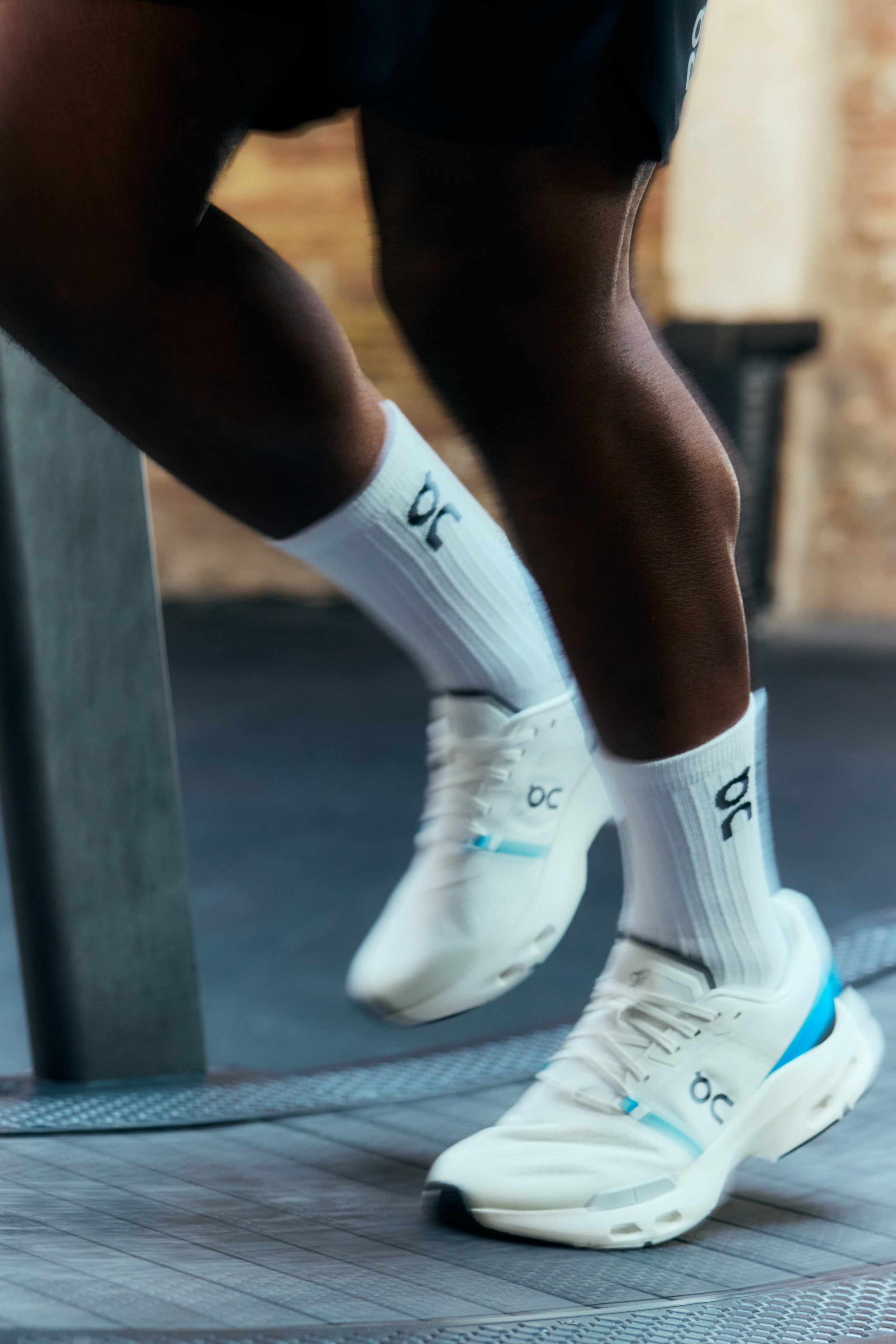Close-up of a person wearing white athletic shoes and socks, stretching on a treadmill with a brick wall background.