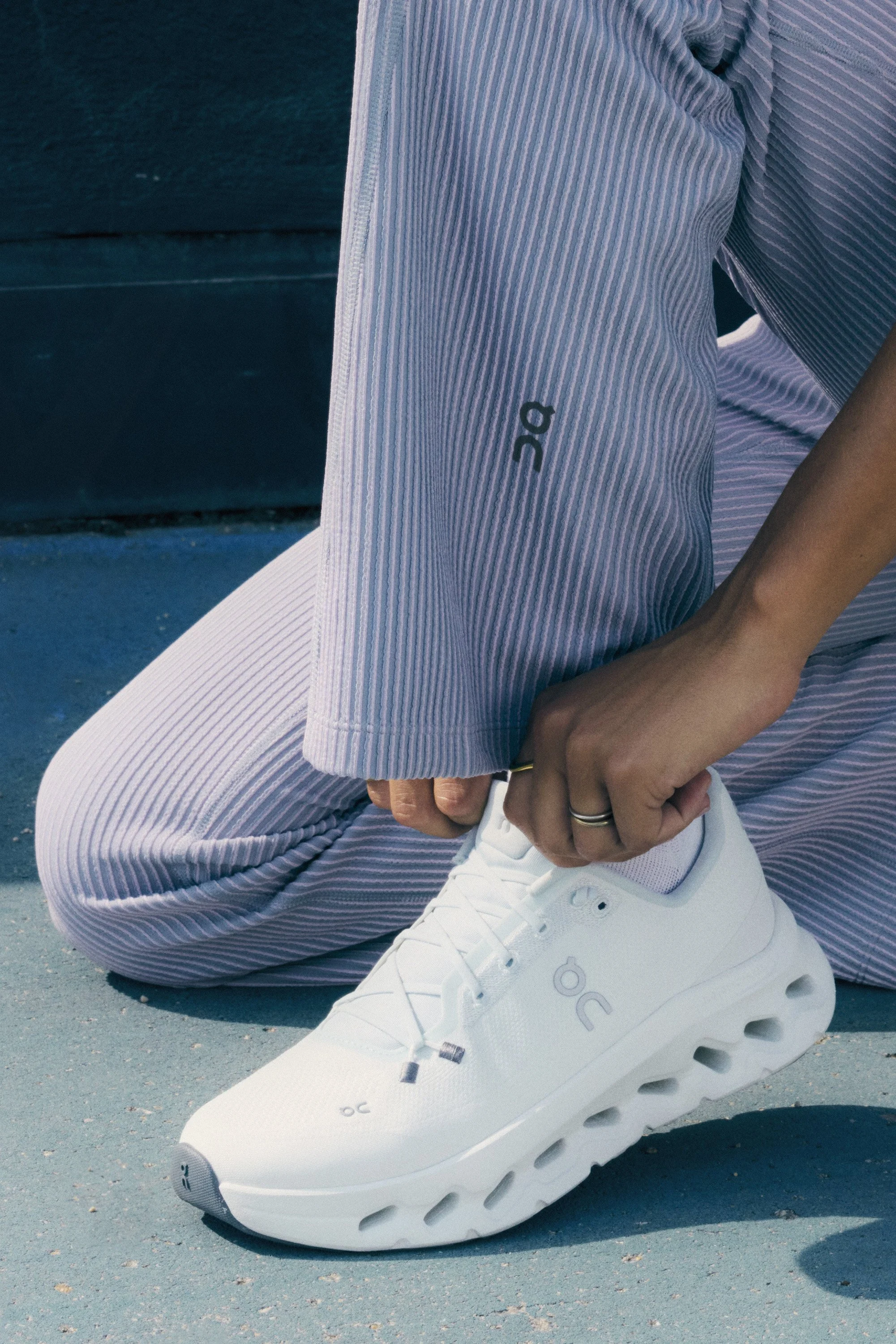 A person in striped athletic wear is kneeling on a blue outdoor court, tying the laces of a white athletic shoe with a unique sole design.