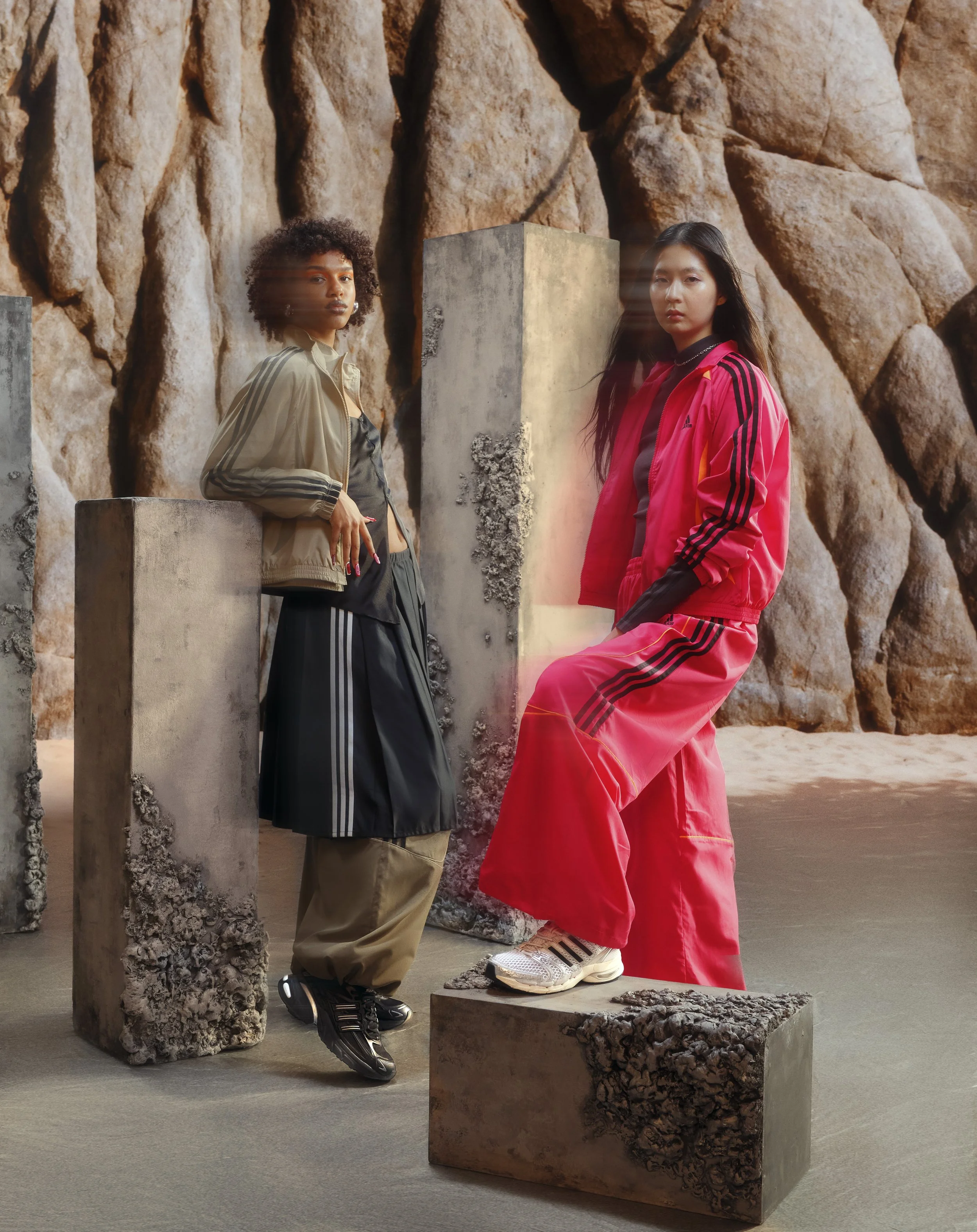 Two women in sporty clothing pose among large concrete blocks with a rocky background, one wearing a beige and black jacket and the other in a red tracksuit.