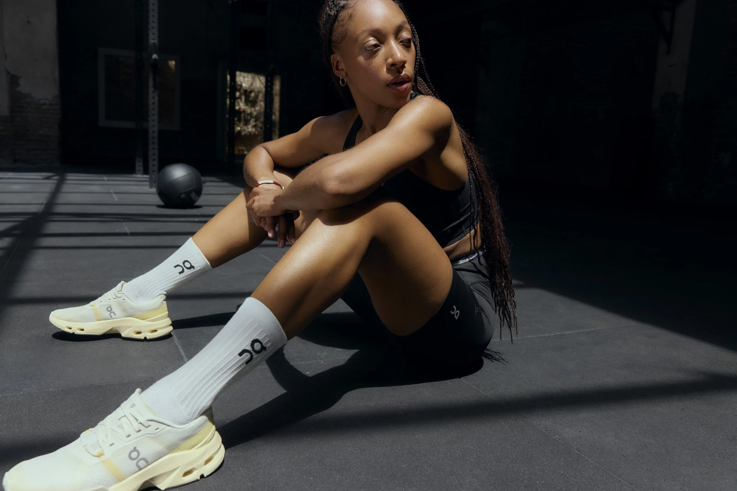 A woman in athletic wear sitting on gym floor, stretching after workout with sunlight and gym equipment in background.