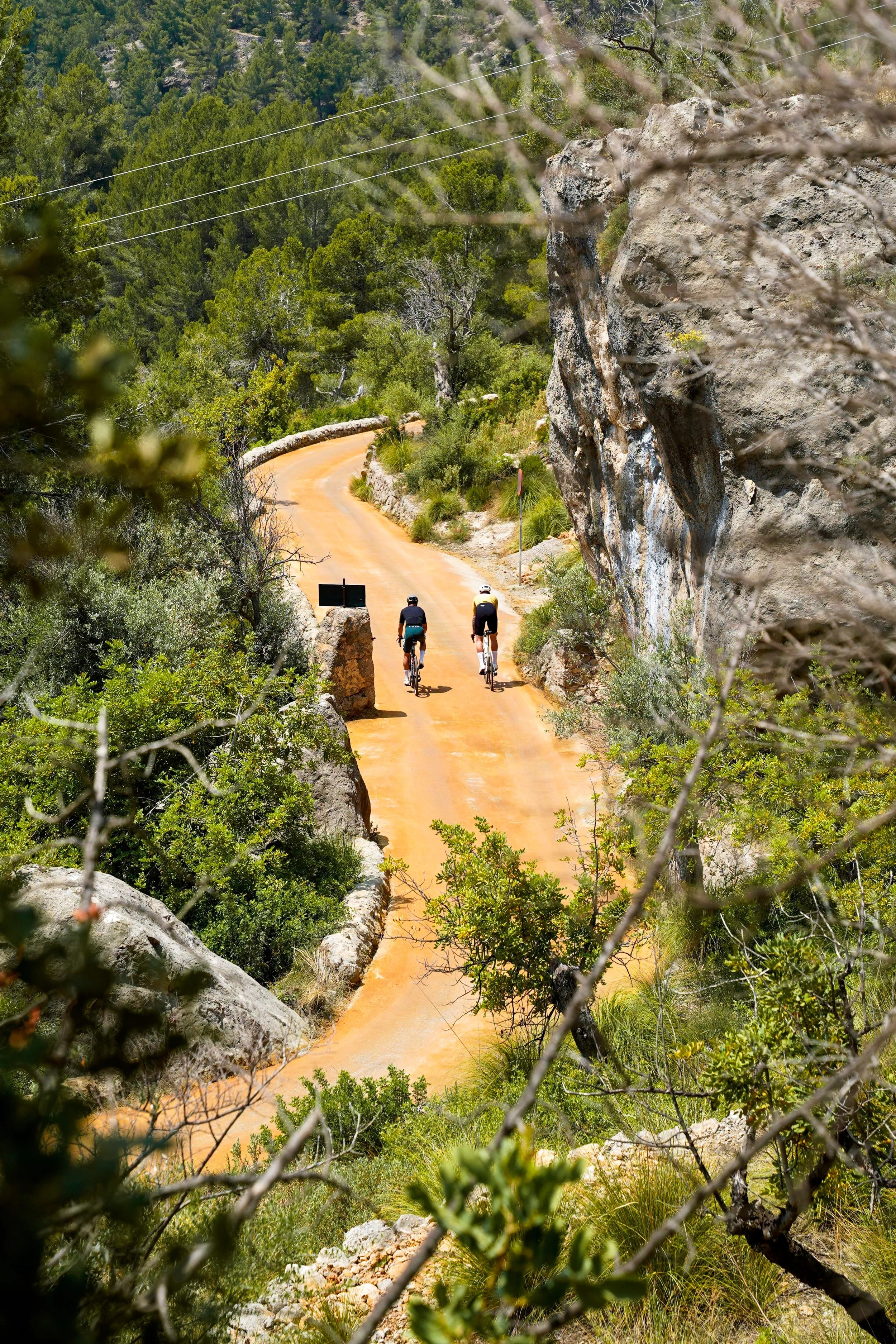 Two cyclists riding on a winding dirt trail through a rocky, forested landscape with trees and shrubs, with large rocks and a cliff on the right side.