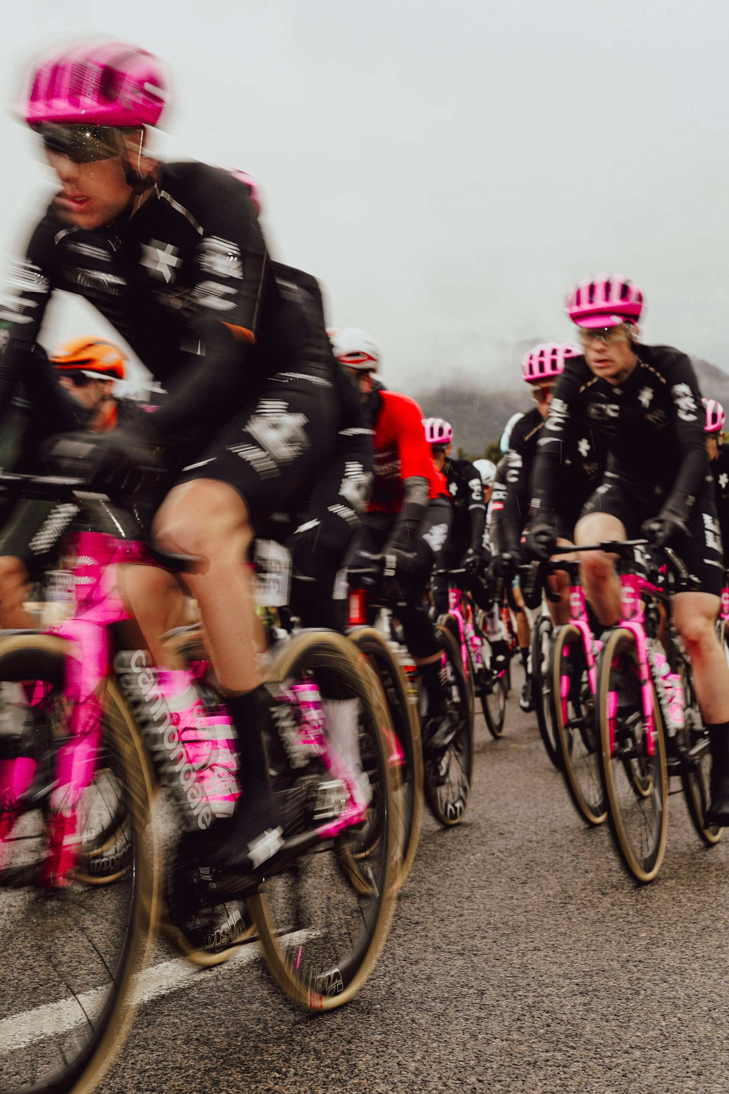 Cyclists participating in a race on a wet, overcast day, wearing black uniforms with pink helmets and pink bikes.