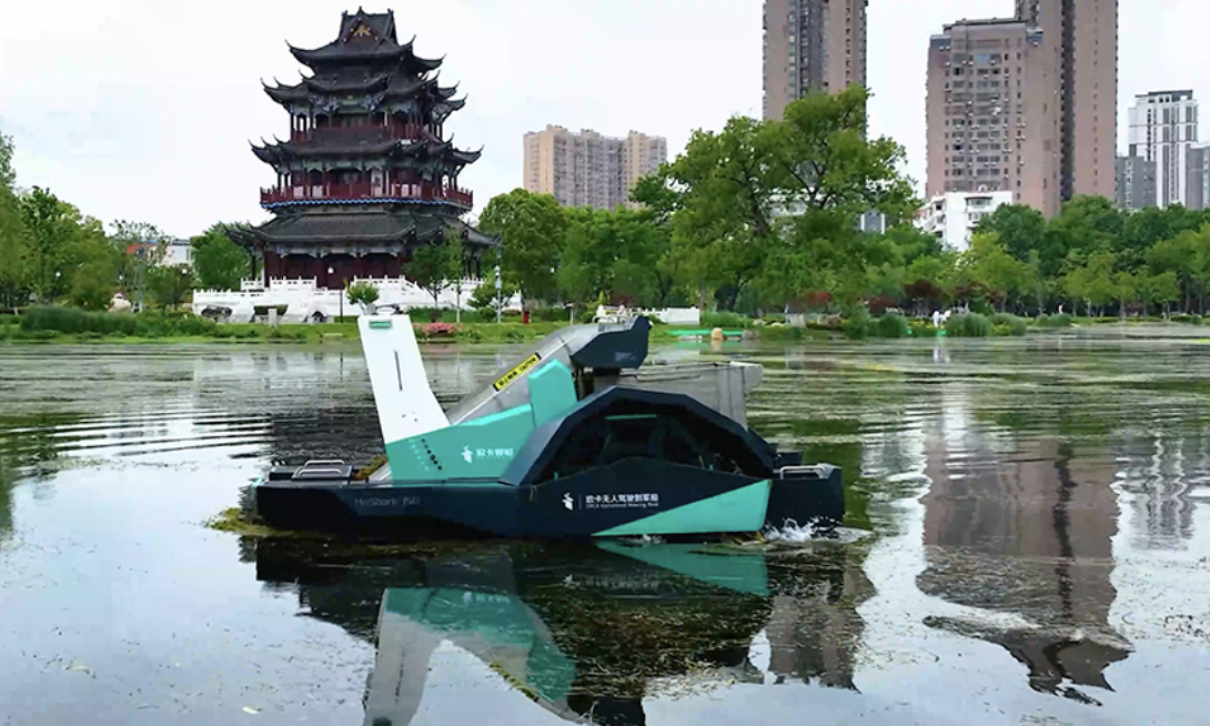 Un bateau nettoyeur de rivière autonavigue sur un lac, avec un pavillon traditionnel chinois en arrière-plan. Des bâtiments modernes et des arbres entourent la scène.