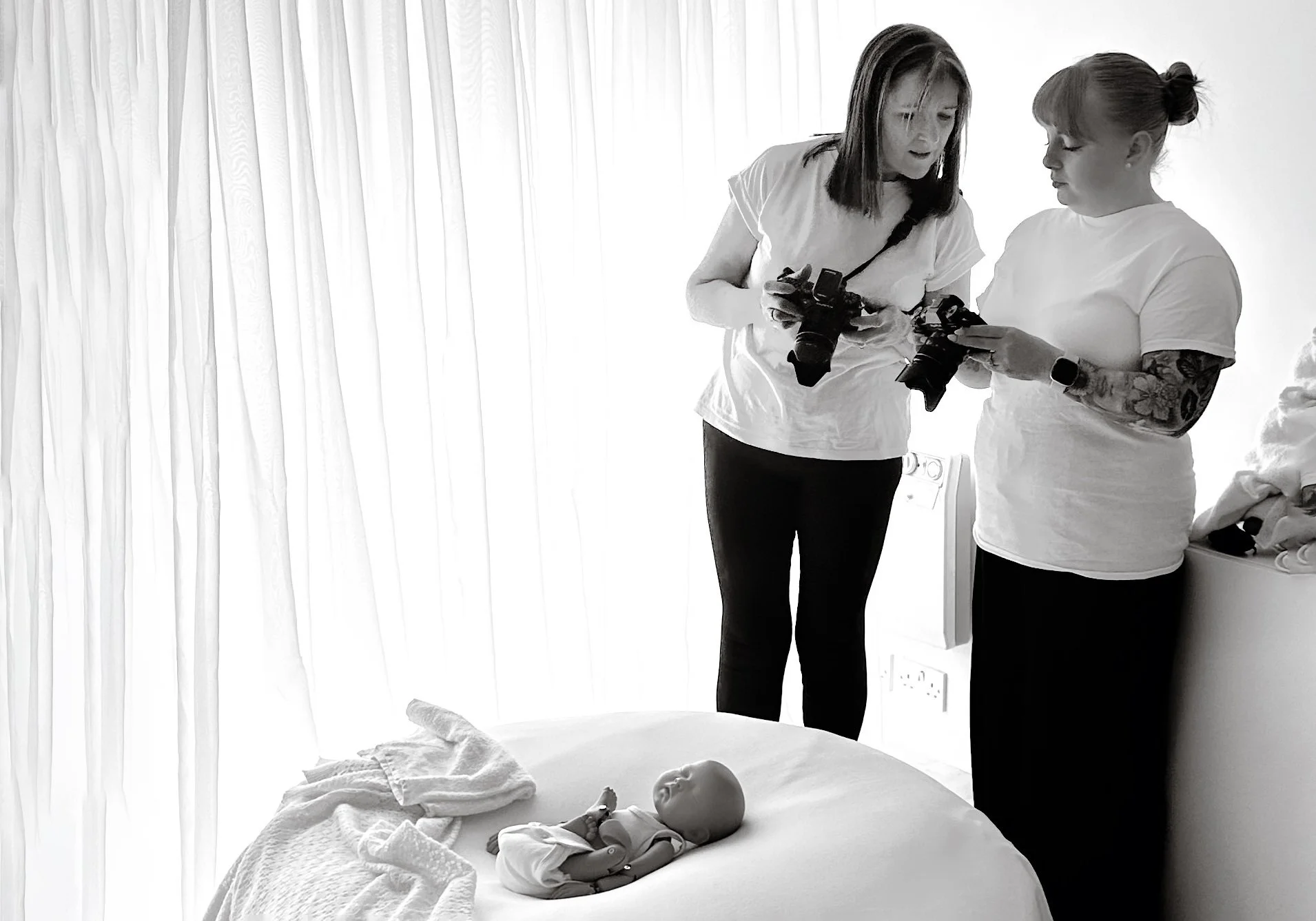 Black and white image of Ally Stuart-Ross guiding a student photographer with cameras during a hands on newborn photography mentoring session with a StandInBaby on a white beanbag