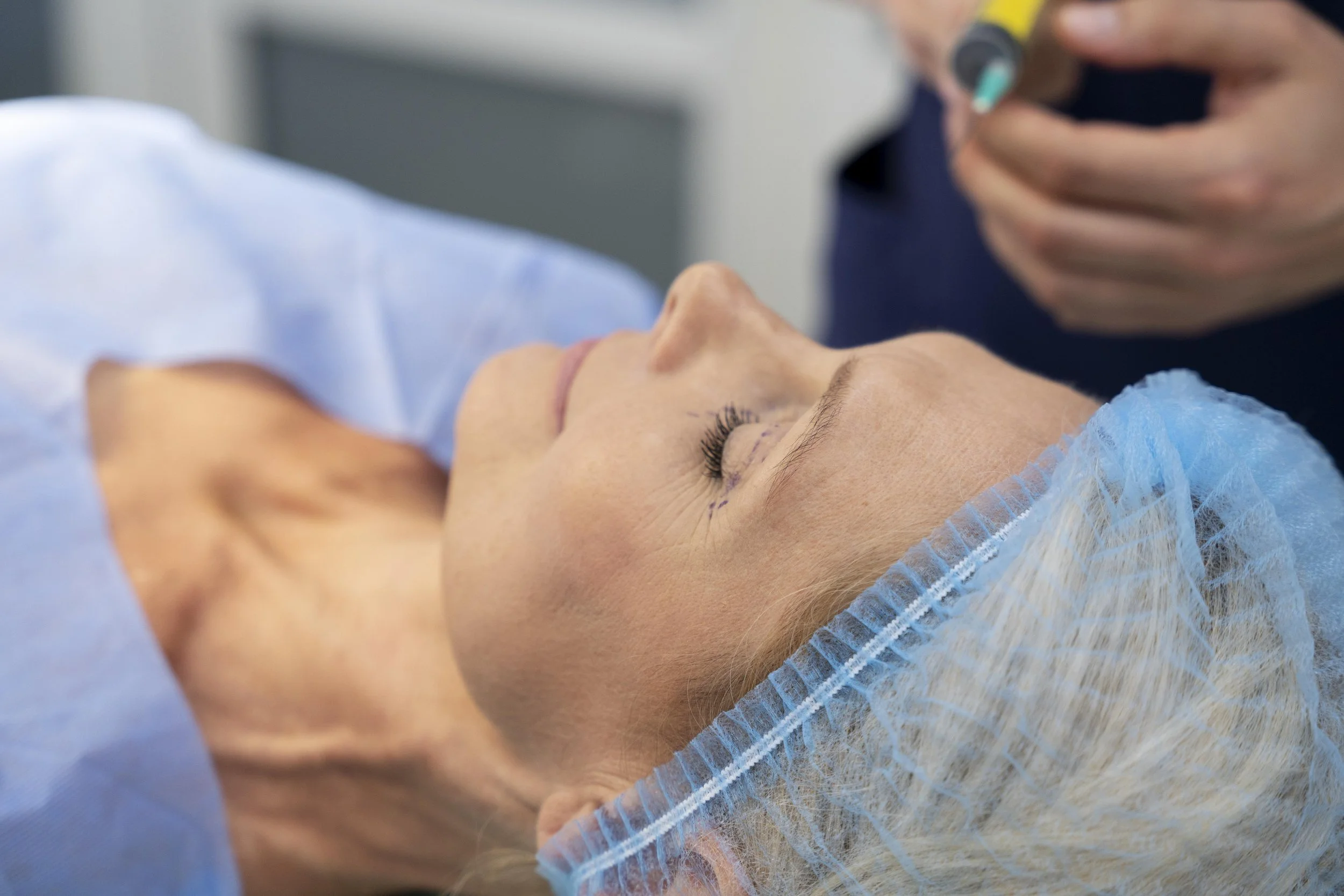 A woman lying on an operating table with eyes closed, wearing a blue surgical cap, with a person nearby holding a medical instrument.