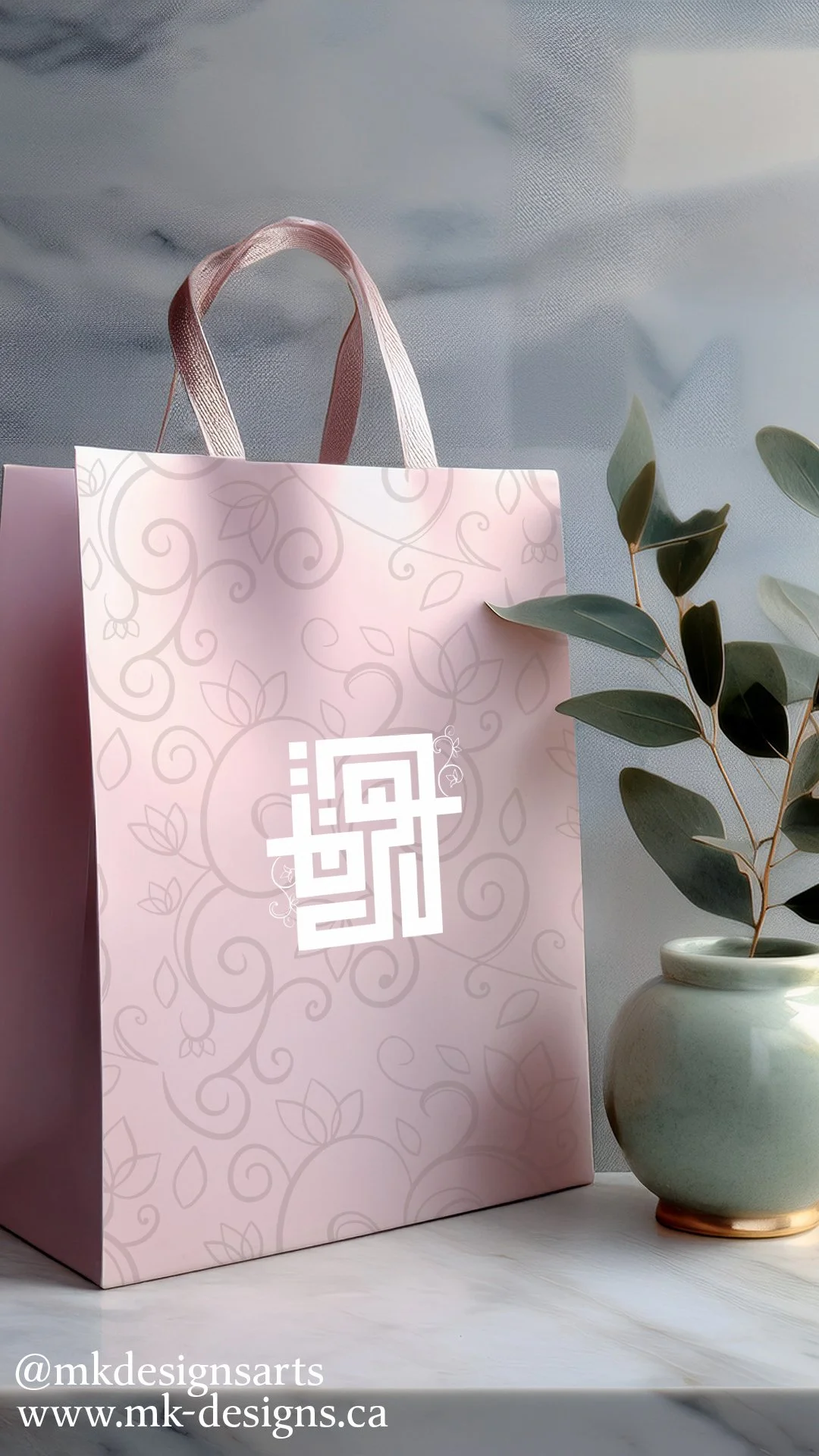 Pink gift bag with white geometric design and logo, next to a white vase with green foliage, on a white surface.
