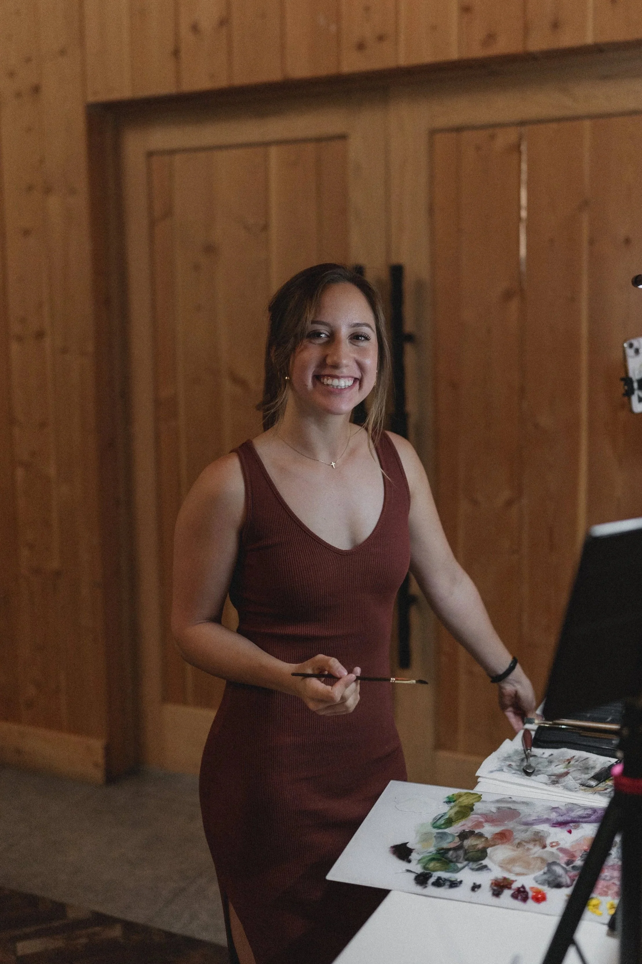 A woman in a sleeveless maroon dress smiling while holding a paintbrush, standing next to a table with a palette of paints and a laptop.