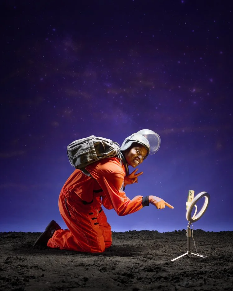 A person in an orange spacesuit and helmet kneeling on the ground at night with a starry sky in the background, pointing at a device on a stand that is emitting light.
