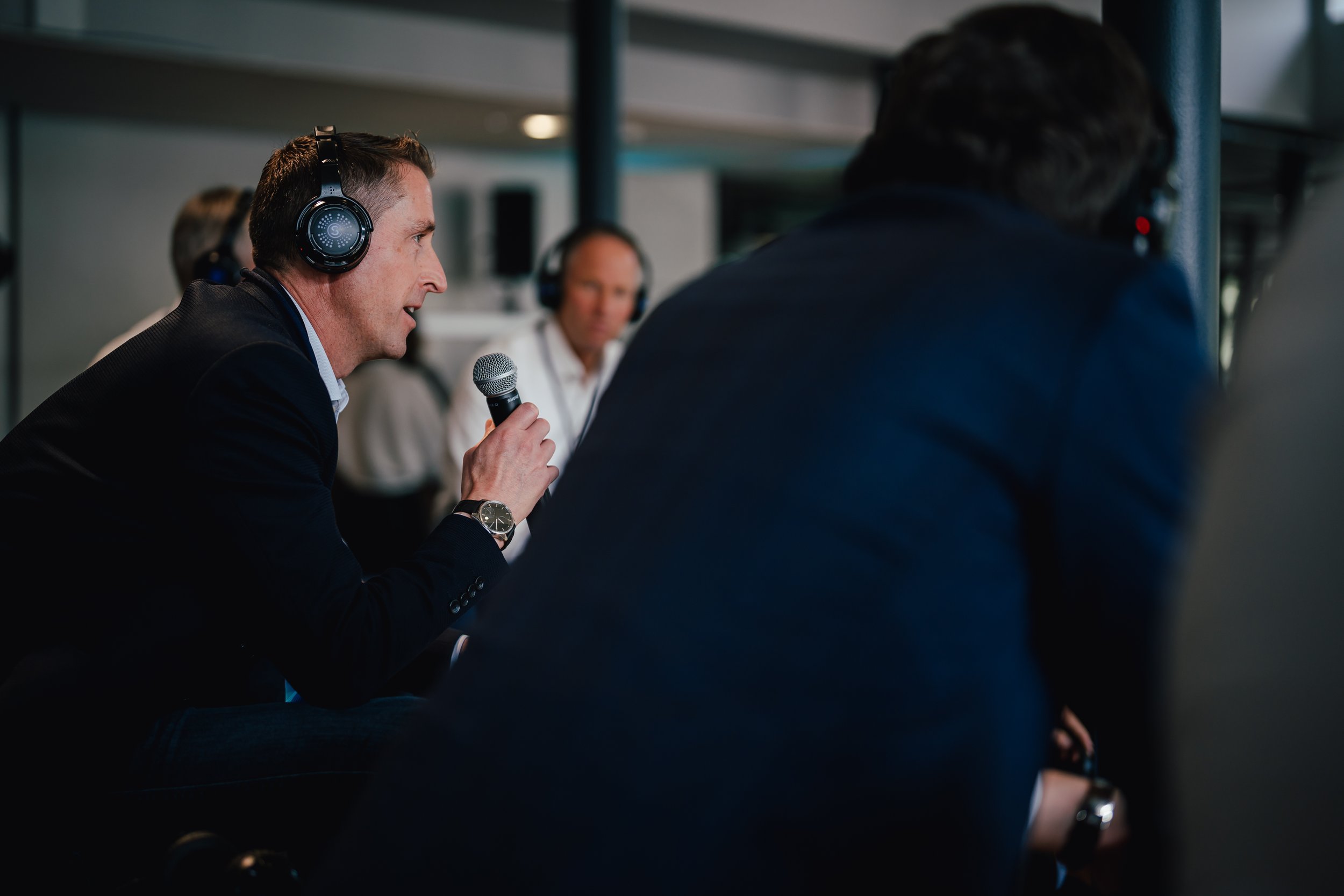 Mann mit Kopfhörern spricht in ein Mikrofon, umgeben von anderen Personen, in einem Konferenzumfeld.