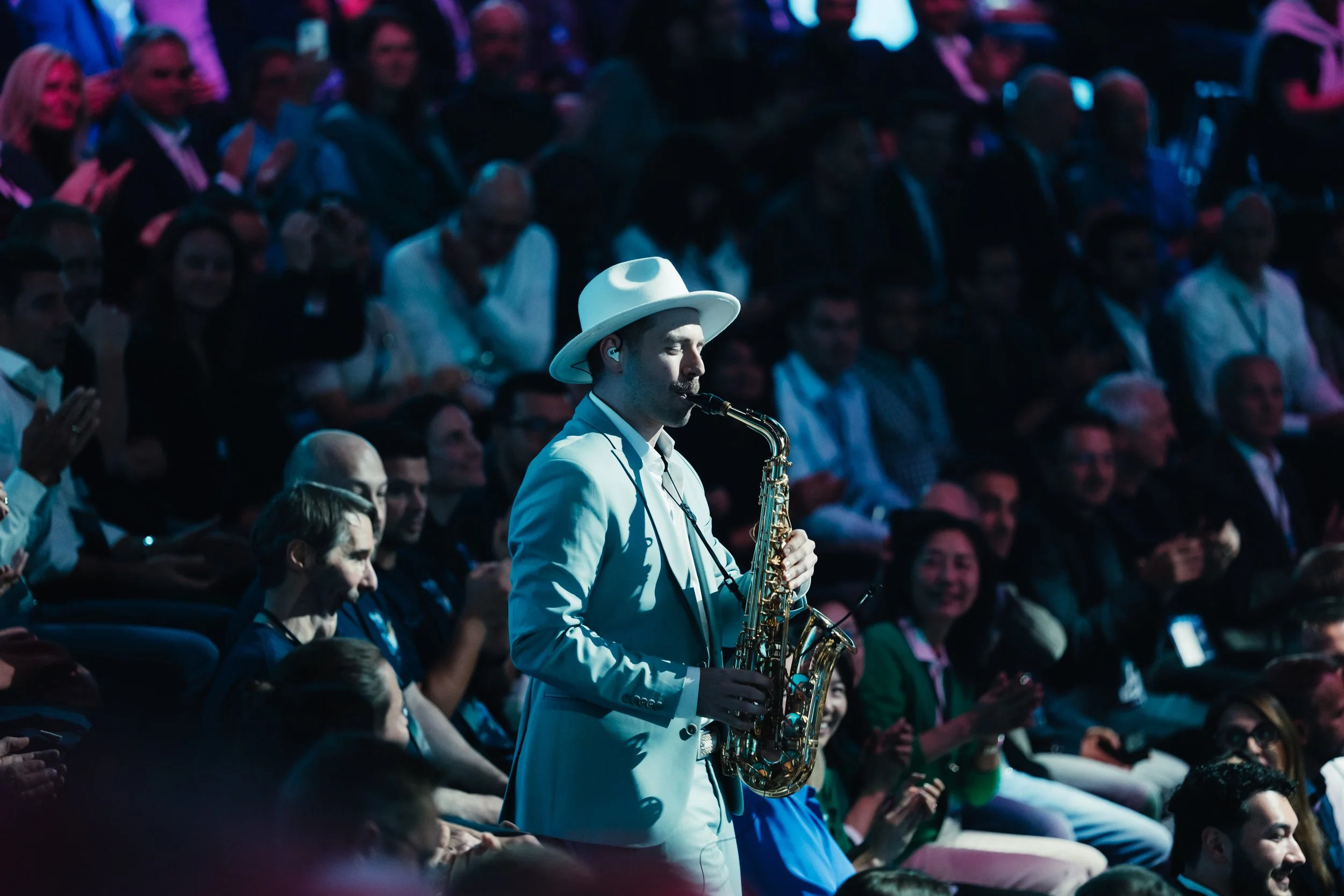 Mann in weißem Anzug und Hut spielt Saxophon vor Publikum bei Konzert.