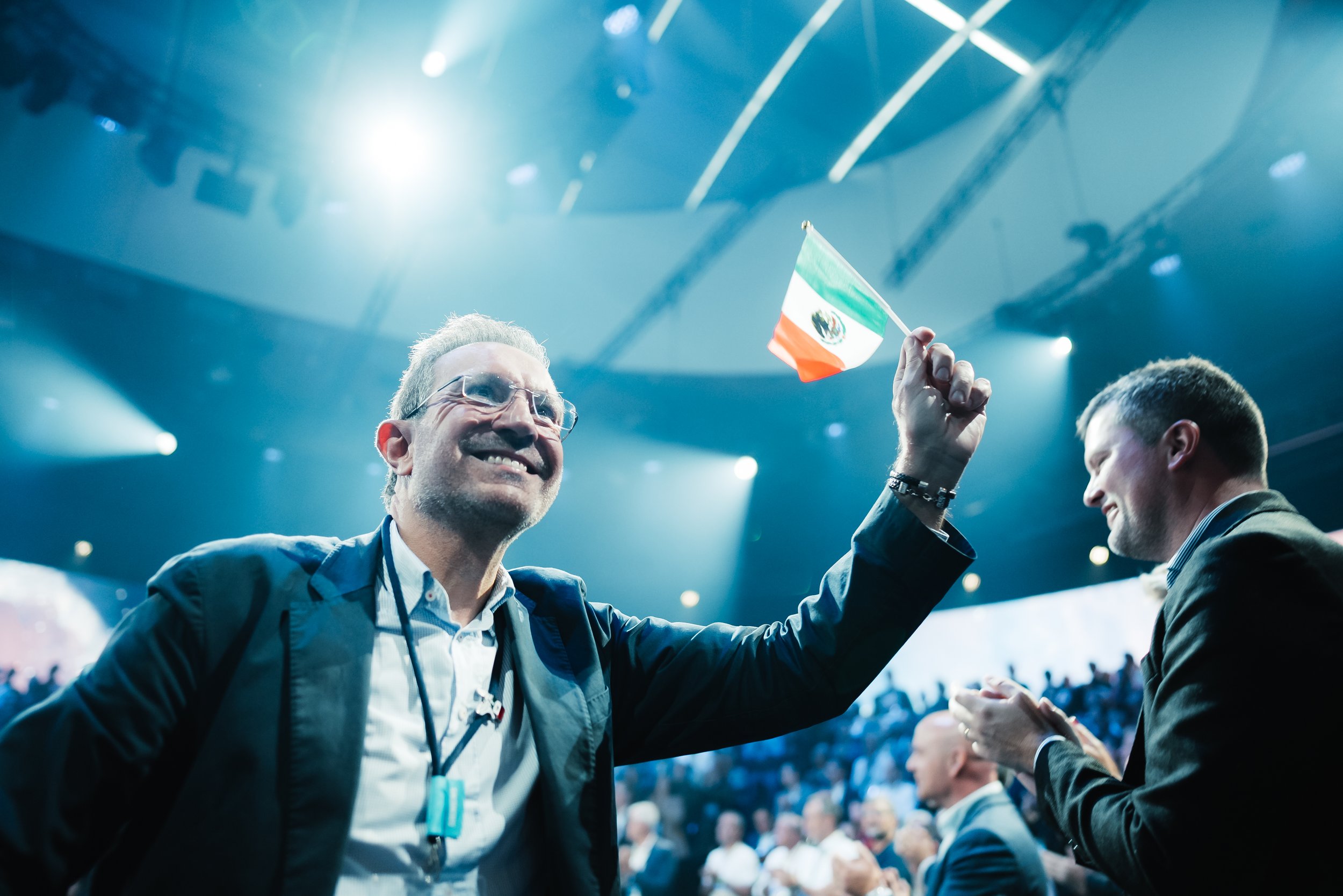 Zwei Männer auf einer Bühne, einer hält eine mexikanische Flagge und lächelt, die andere applaudiert.