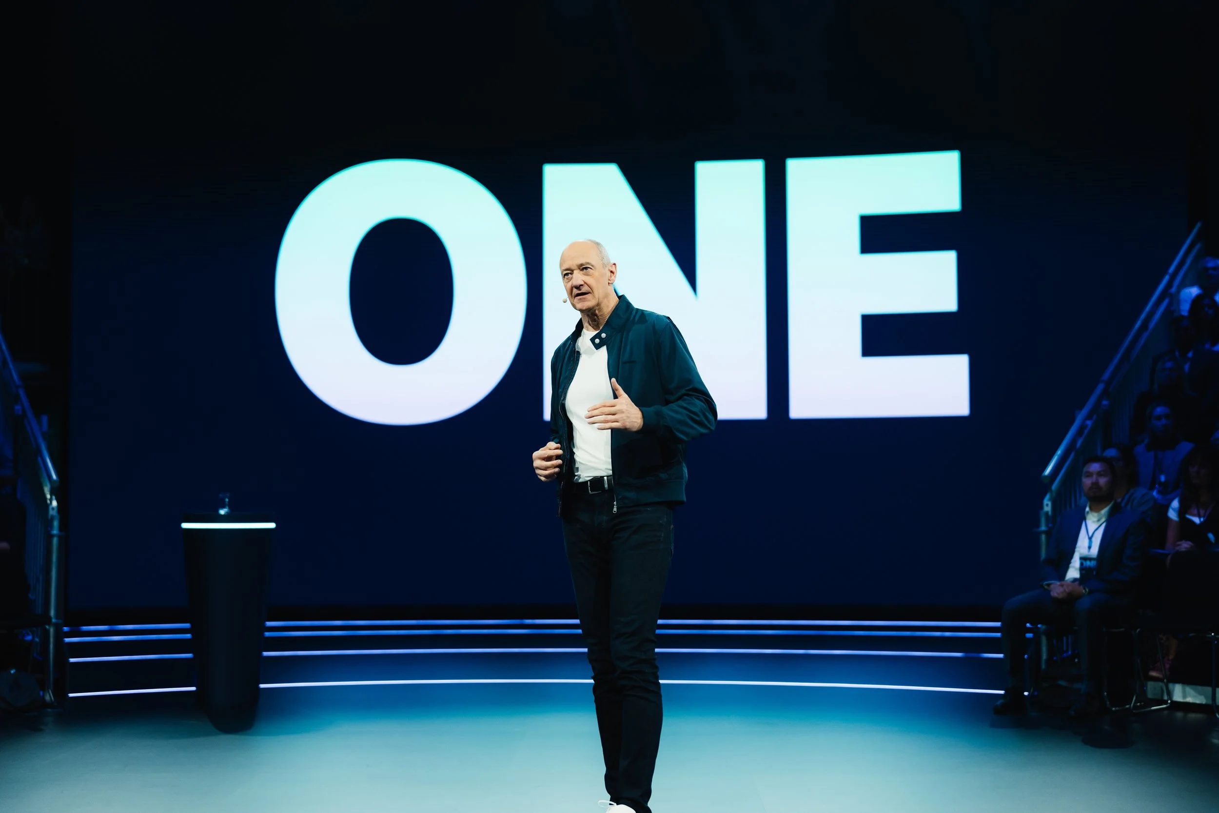 Ein Mann spricht auf einer Bühne vor einem großen Bildschirm mit der Aufschrift 'ONE'.