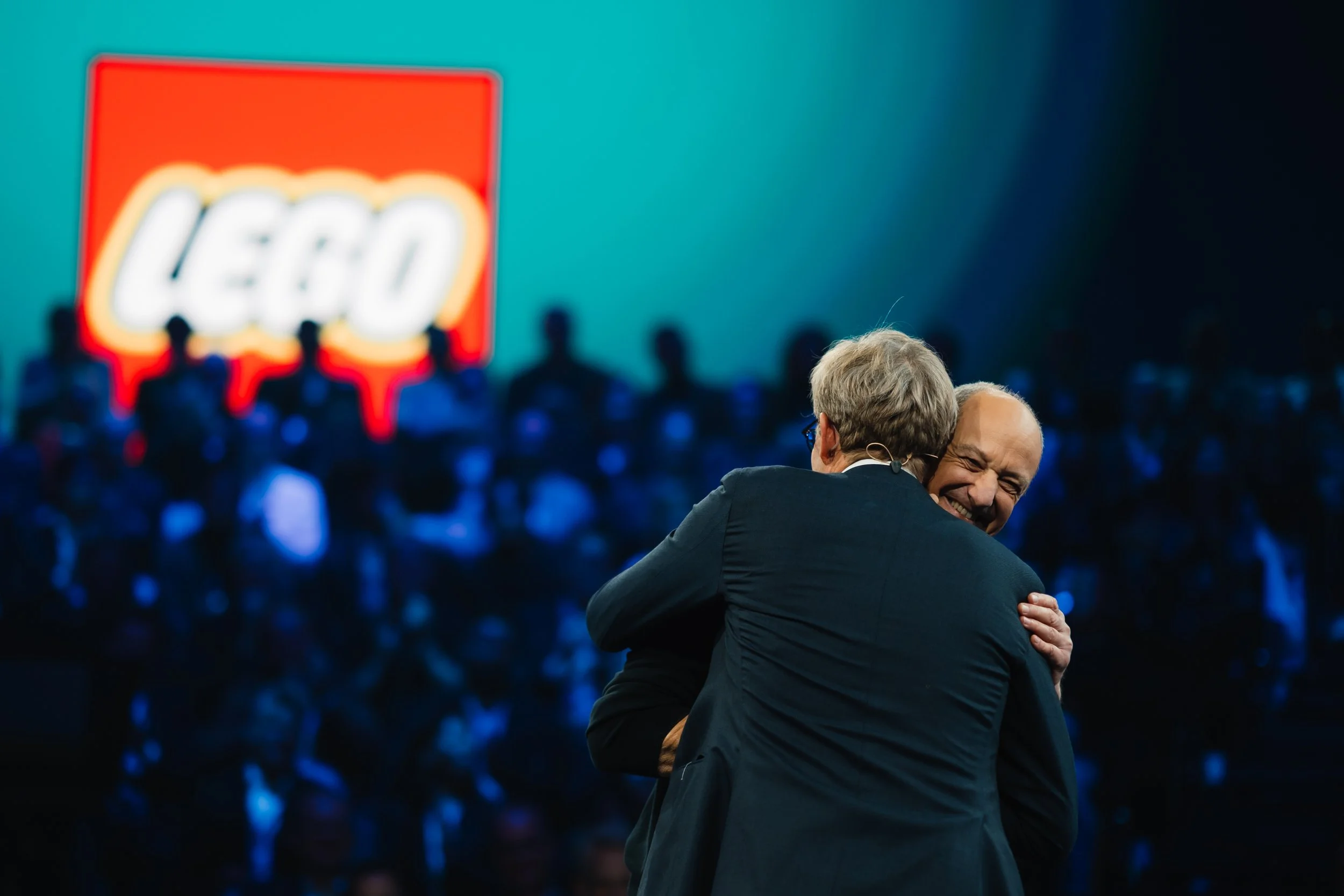 Zwei Männer umarmen sich auf einer Bühne, im Hintergrund ein großes LEGO-Logo und ein Publikum.