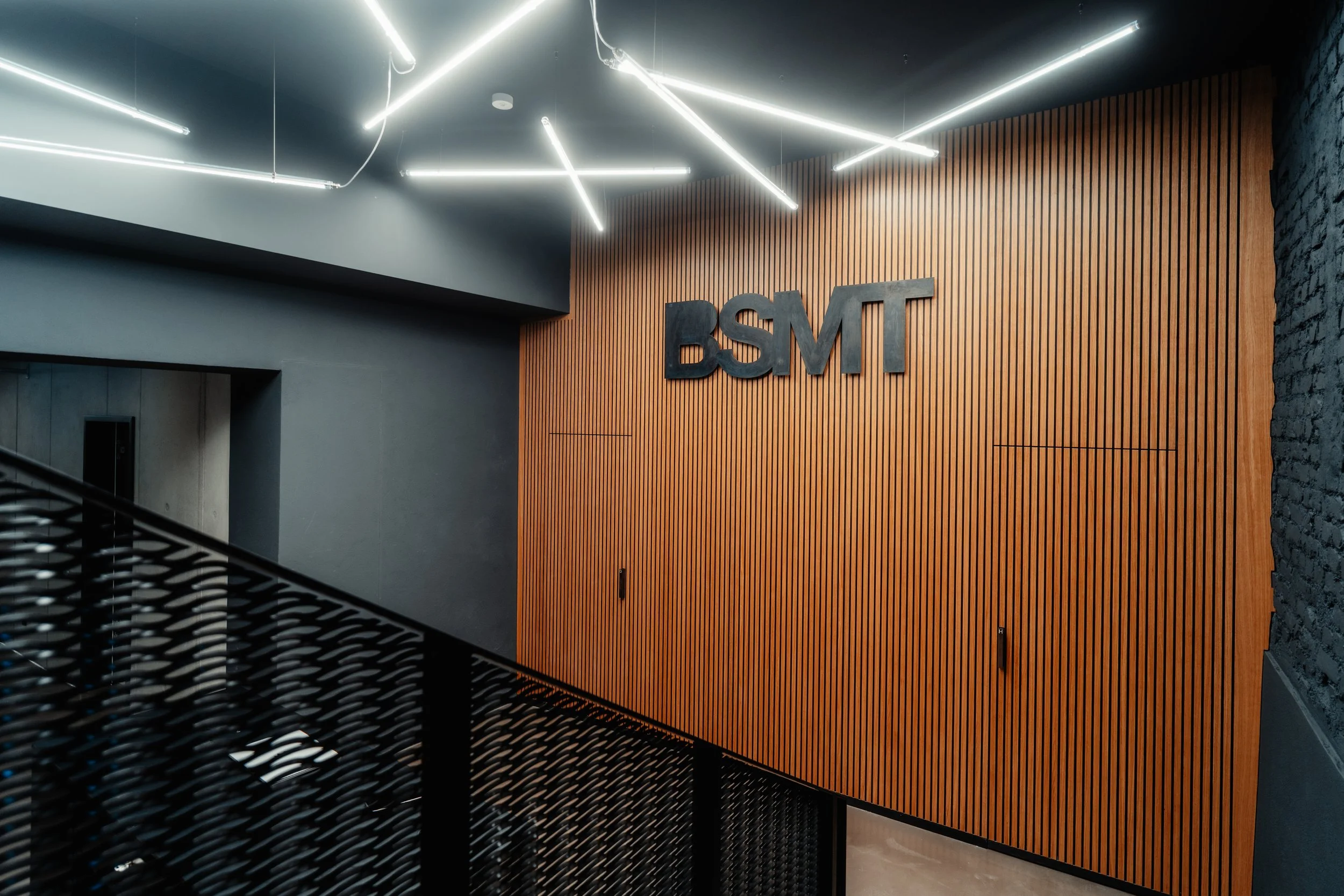 Innenansicht eines modernen Gebäudes mit einer Holzverkleidung, einer dunklen Wand und einer Galerie mit schwarzen Metallgeländer, beleuchtet durch weiße LED-Leuchten.