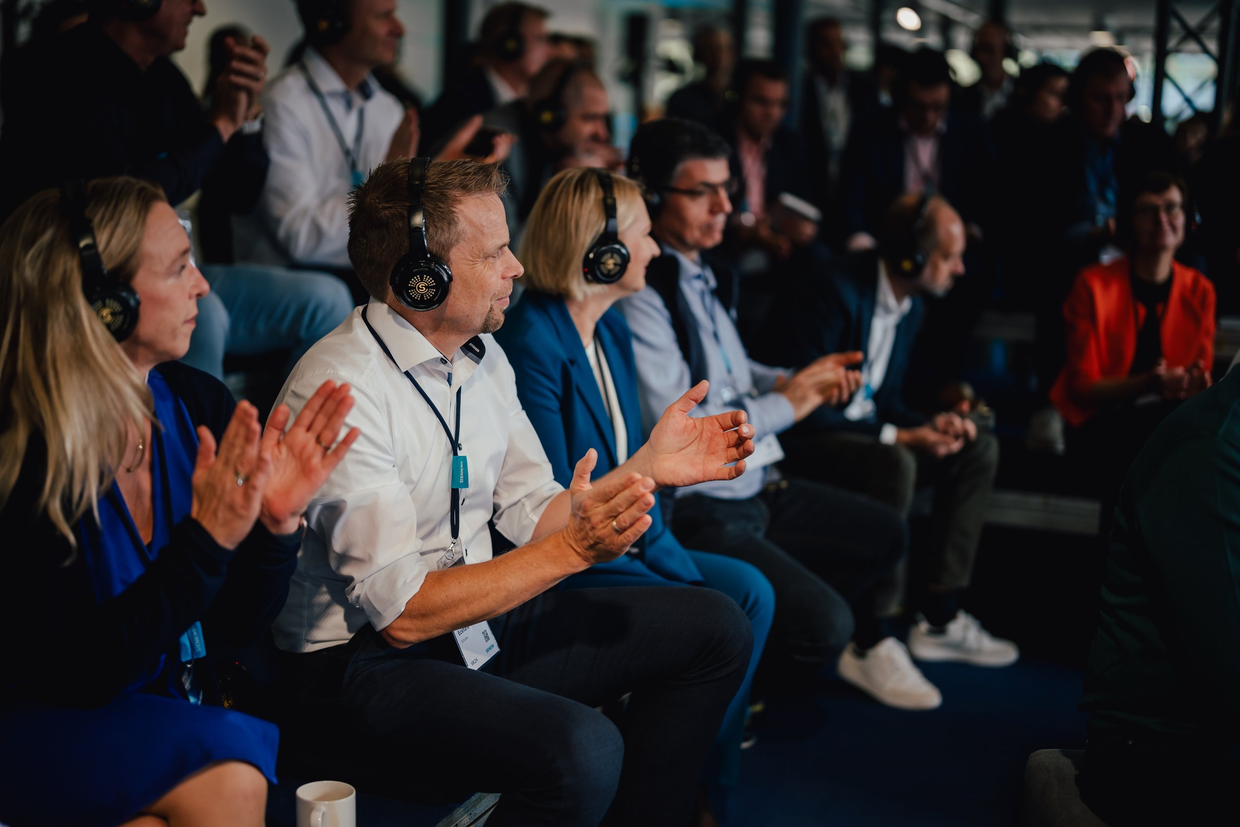 Gruppe von Personen sitzt in einem Konferenzraum, trägt Kopfhörer und applaudiert, teilweise Business-Kleidung, konzentriert auf eine Präsentation oder Veranstaltung.