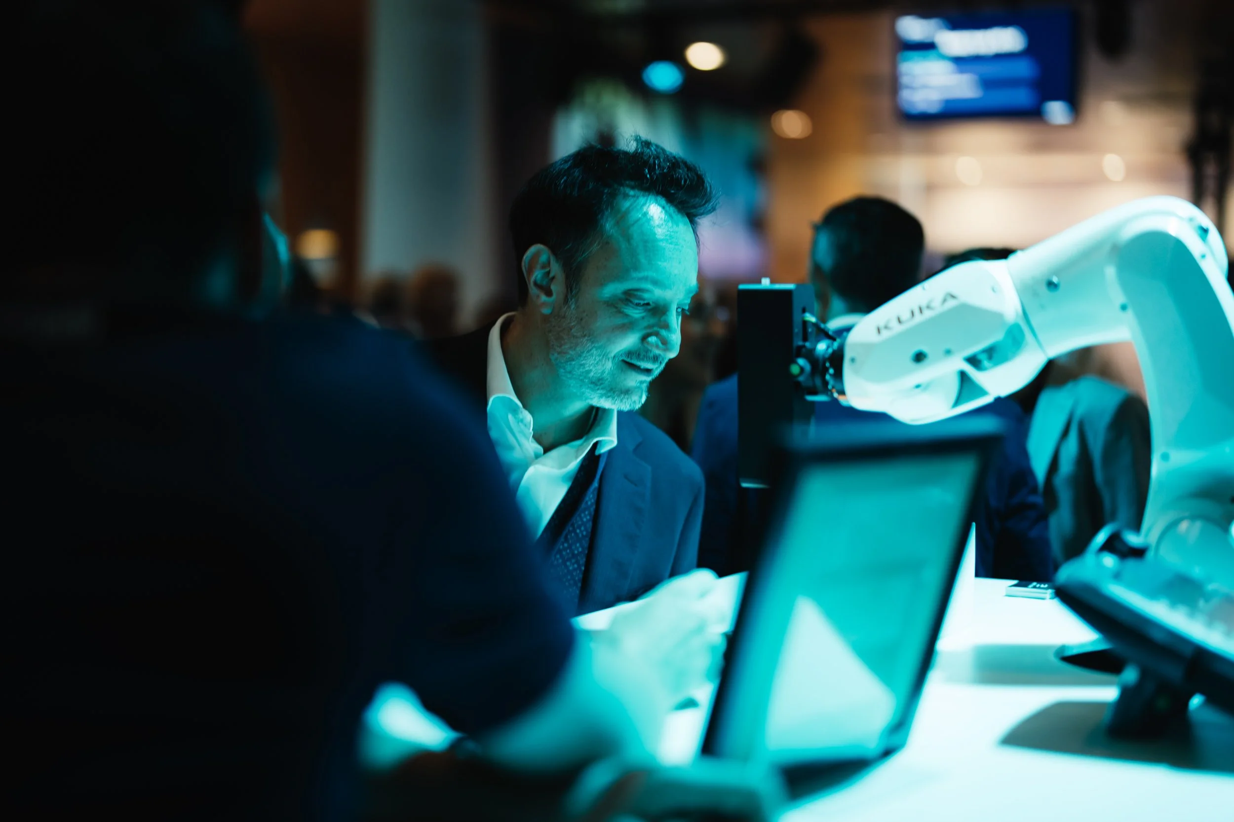 Ein Mann betrachtet ein Objekt, das von einem Roboterarm mit der Aufschrift 'KUKA' gehalten wird, in einem modernen, gut beleuchteten Raum mit mehreren anderen Menschen im Hintergrund.