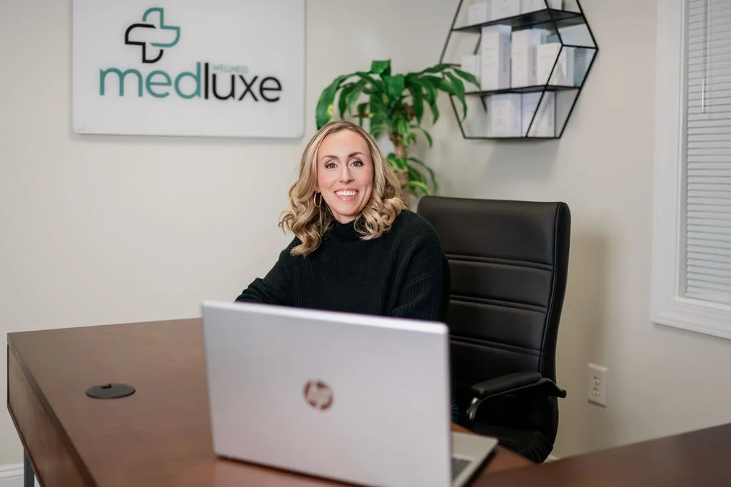 A woman sitting at a desk in an office, smiling, with a laptop in front of her. Behind her is a sign that says "medluxe" with a cross logo, and a green potted plant. There is a black geometric shelf with documents or books on the wall to her right.
