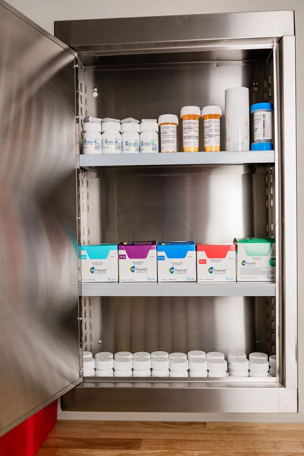 A stainless steel medicine cabinet with multiple shelves containing prescription medication bottles and boxes, some with white caps and labels.