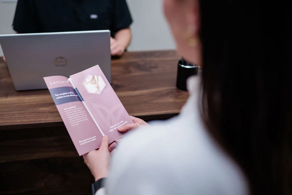 A woman reading a pink brochure while sitting at a wooden table in an office, with a man working on a laptop across from her.