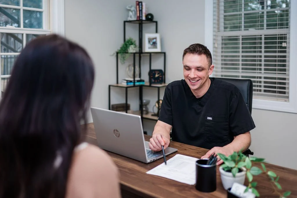 A male healthcare professional smiling at a woman during an appointment in an office with a desk, computer, and shelf with decor in the background.