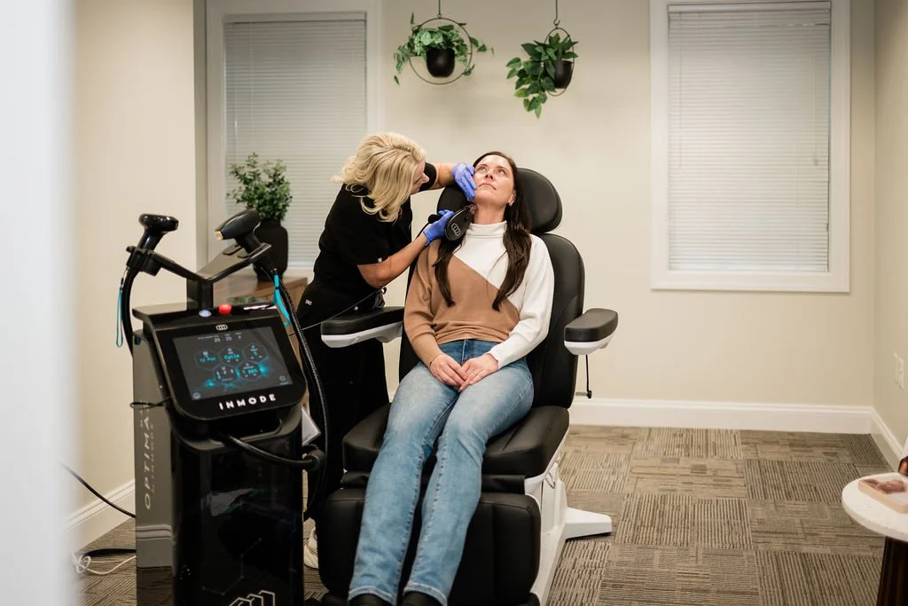 A woman receiving a cosmetic or skincare treatment from a professional in a medical office or clinic. The woman is seated in a black reclining chair, while the professional, wearing gloves, is performing a procedure near her face. The room has a modern appearance with white walls, two windows with white blinds, and hanging plants.
