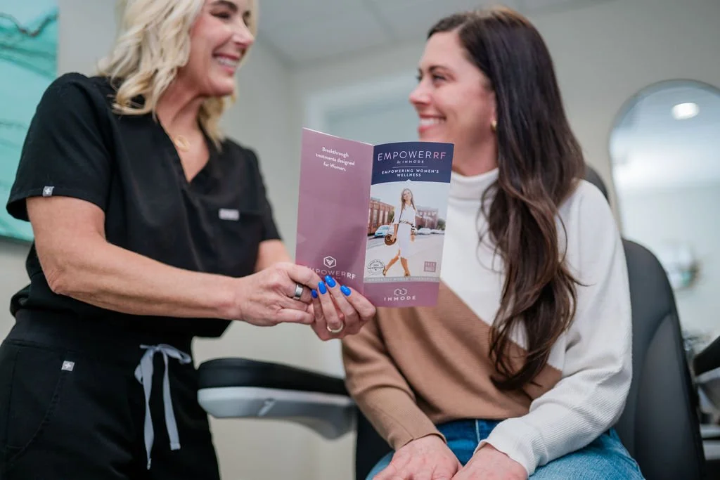 A woman in a black uniform showing a pamphlet to another woman with long brown hair sitting in a chair, inside a professional office setting.