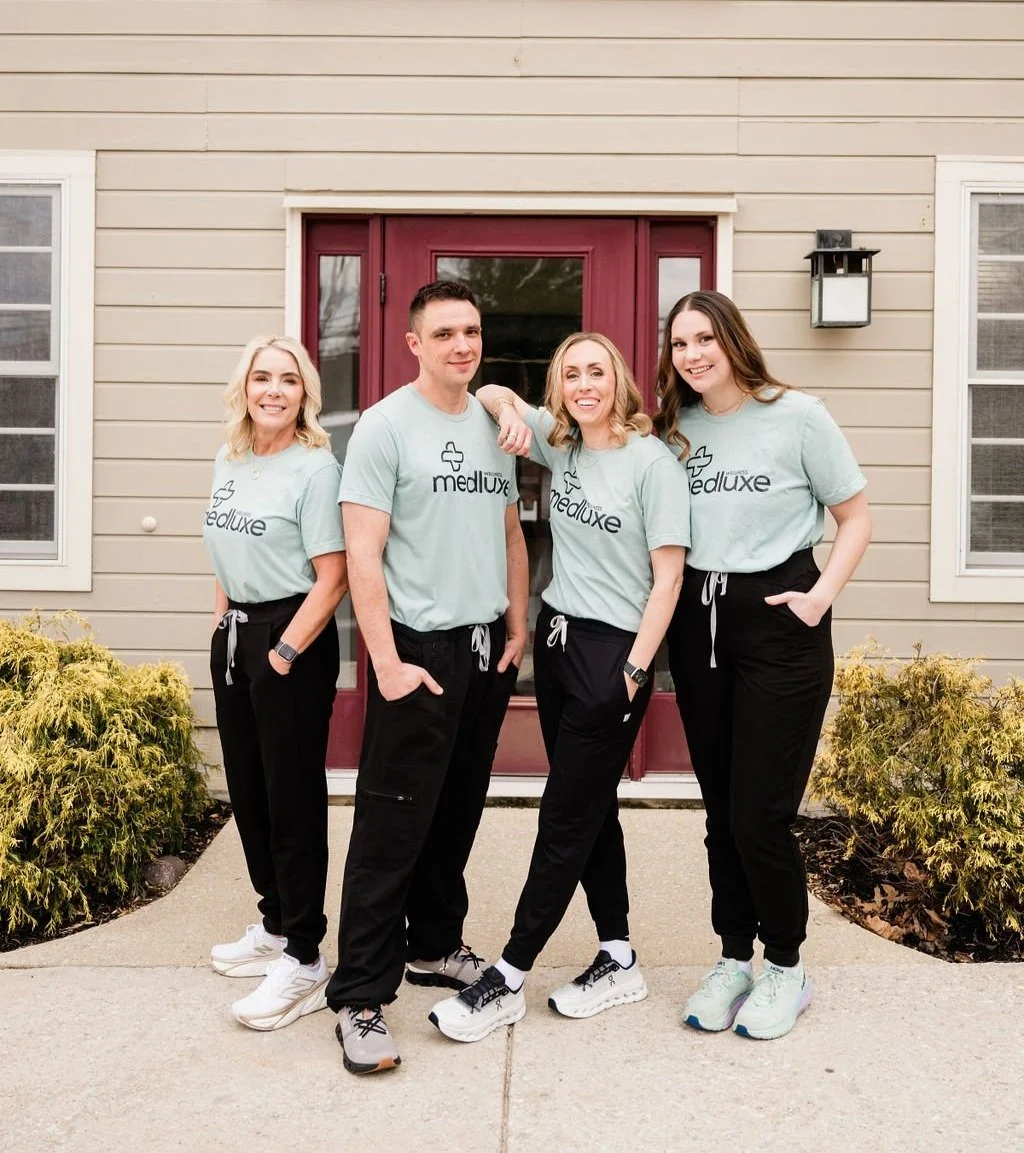 Four people standing outside in front of a beige house with a maroon door, wearing matching light blue Medluxe shirts and black pants, smiling at the camera.