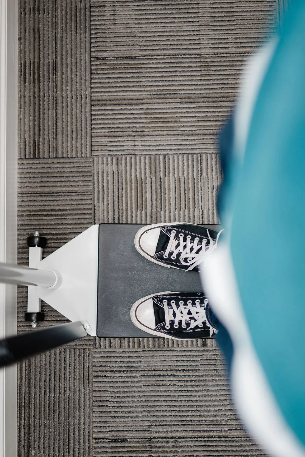 Top-down view of a person in black sneakers standing on a gray chair mat on a carpeted floor, with a blue garment partially visible on the right.