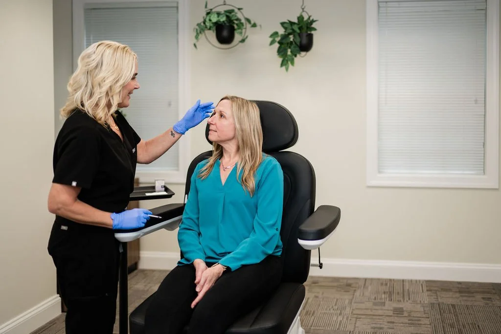 A woman doctor in black scrubs and blue gloves examining a woman's nose or face in a medical office.