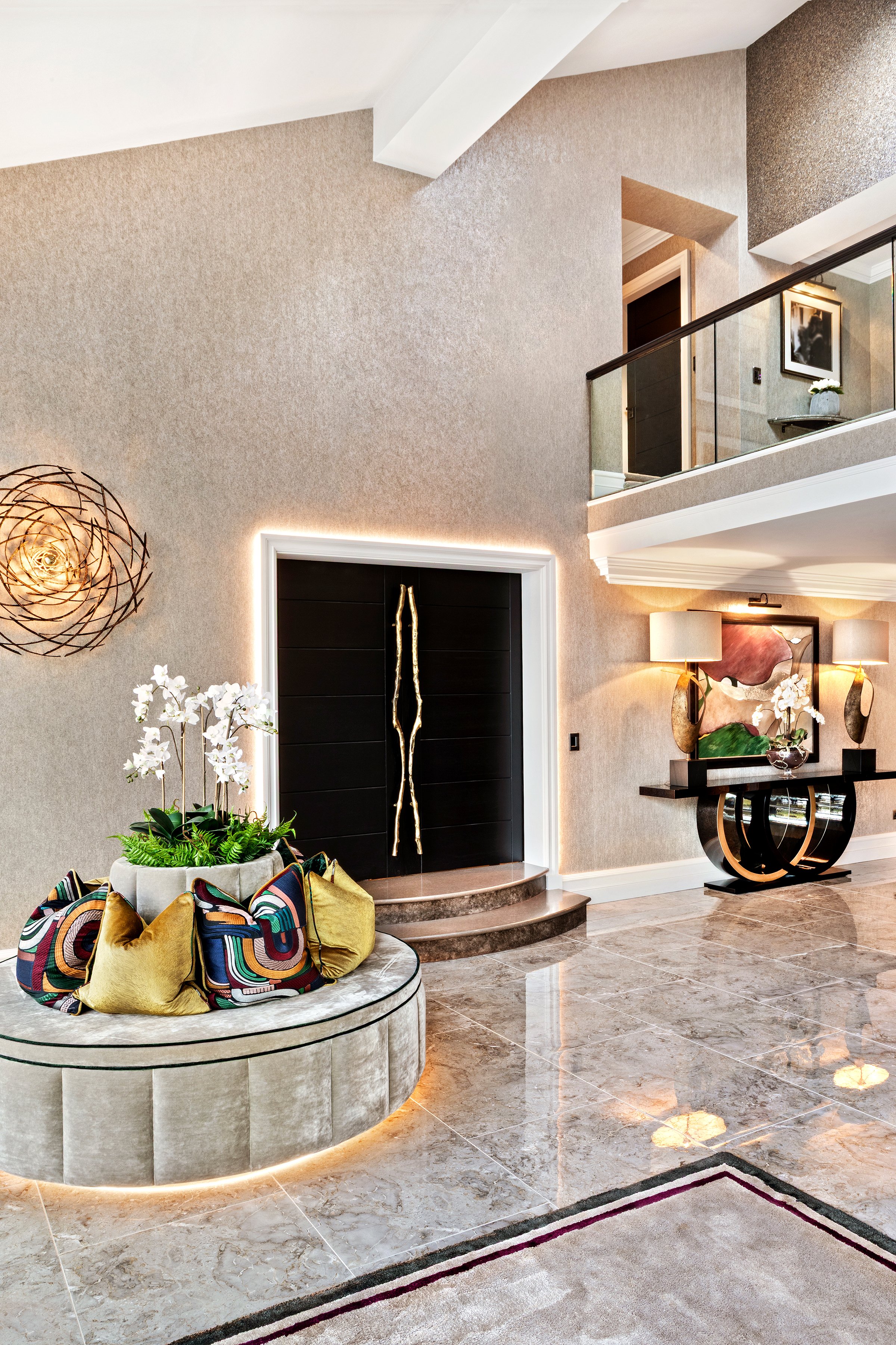 Interior of a modern house with a beige textured wall, stylish black front door with gold handles, a round velvet seating area decorated with colorful pillows and a flower arrangement, marble tile flooring, a staircase leading to an upper level, contemporary artwork and lighting fixtures.