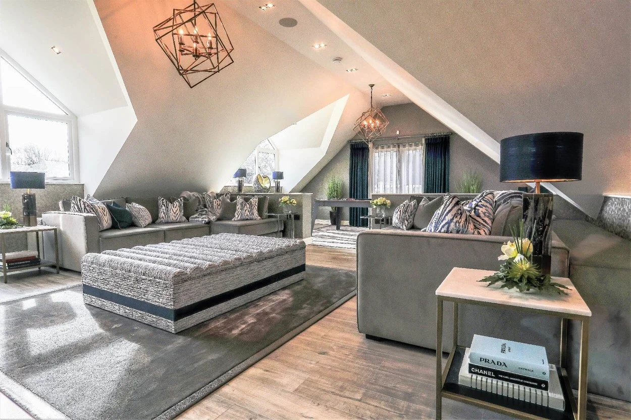 Modern living room with sloped ceiling, featuring two large light gray sofas with patterned pillows, a textured ottoman, and decorative accessories including lamps and books. Large windows with curtains allow natural light, enhancing the neutral colo