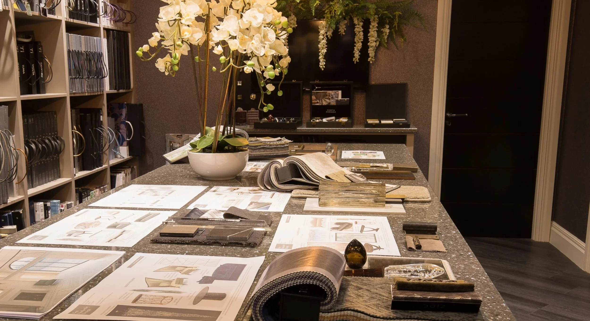 Interior of a design or furniture showroom with a large display table covered in fabric and material swatches, color charts, and design samples. There is a white orchid plant and flowers on the table, and shelves filled with books and color chips along the wall. The background includes a countertop with jewelry or accessory boxes and a sleek black door.