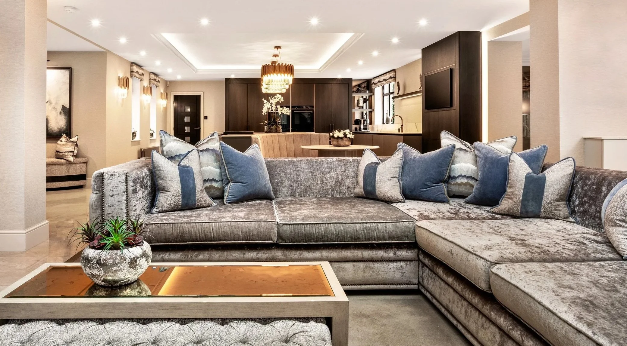 Modern living room with light-colored walls, plush velvet sectional sofa with multiple throw pillows, a rectangular glass-top coffee table with a decorative plant, and dark wood accents including a large kitchen island, cabinets, and a flat-screen TV on the wall. The ceiling has recessed lighting and a central decorative chandelier.