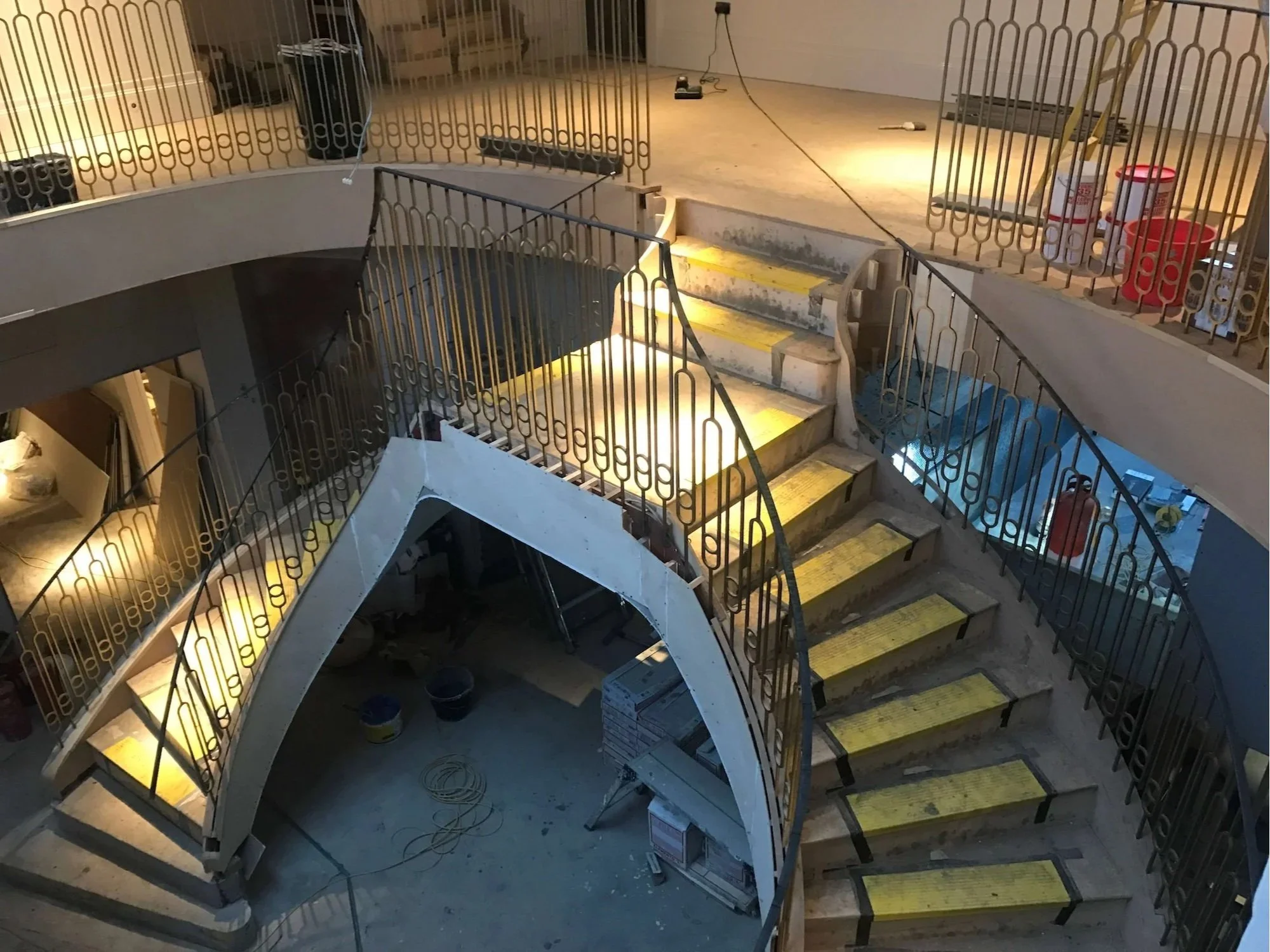 Interior of a building under construction or renovation featuring a staircase with yellow safety strips, temporary fencing or barriers, construction tools, materials, and scattered supplies.