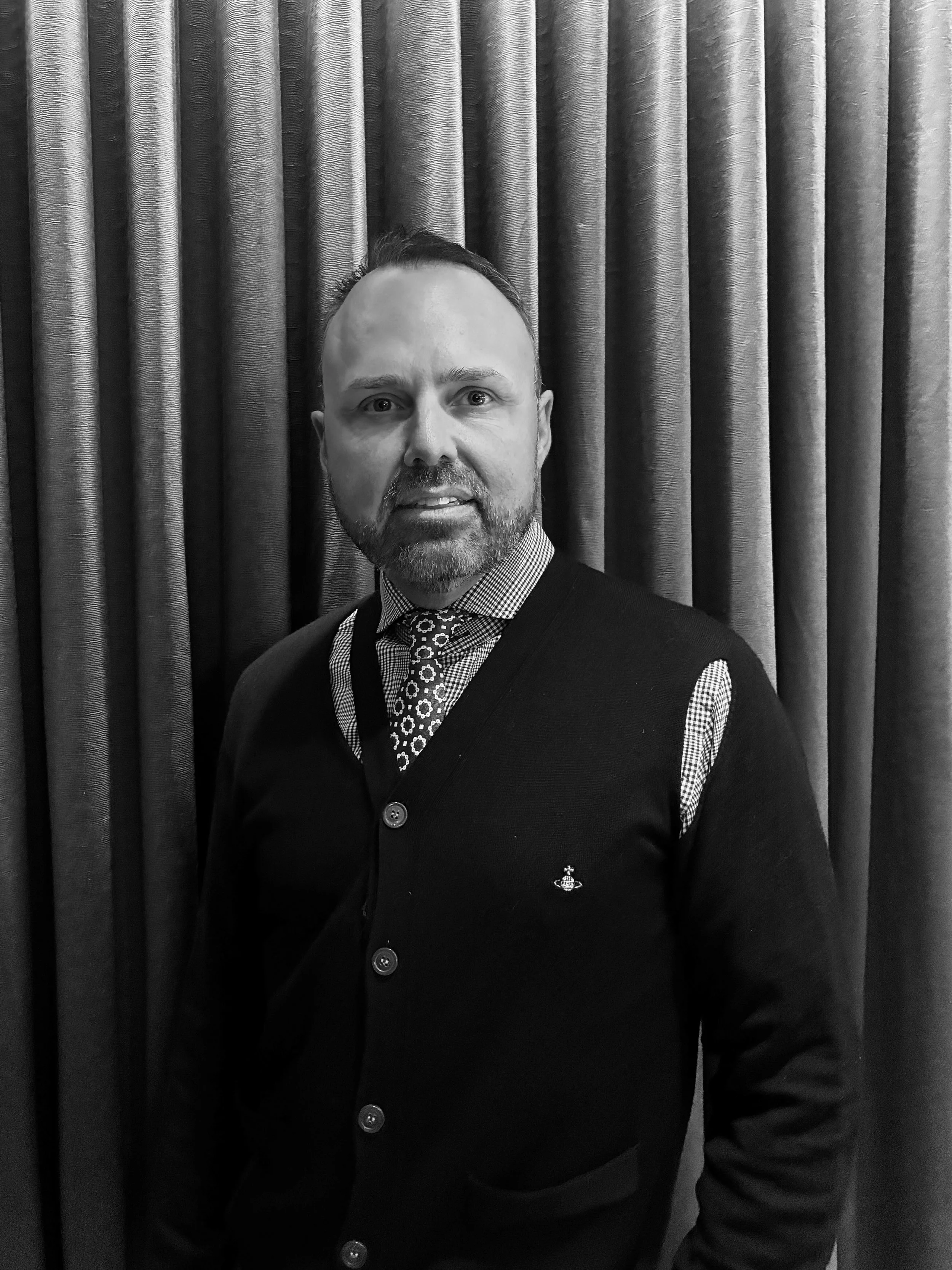 A man with a beard and mustache, wearing a checkered shirt, patterned tie, and dark vest, standing in front of a curtain.
