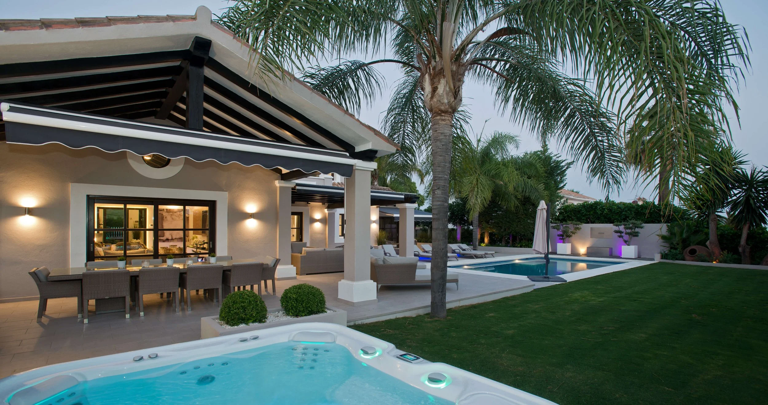Backyard of a house at dusk with a swimming pool, outdoor seating area, palm trees, and landscaped garden, illuminated with exterior lights.