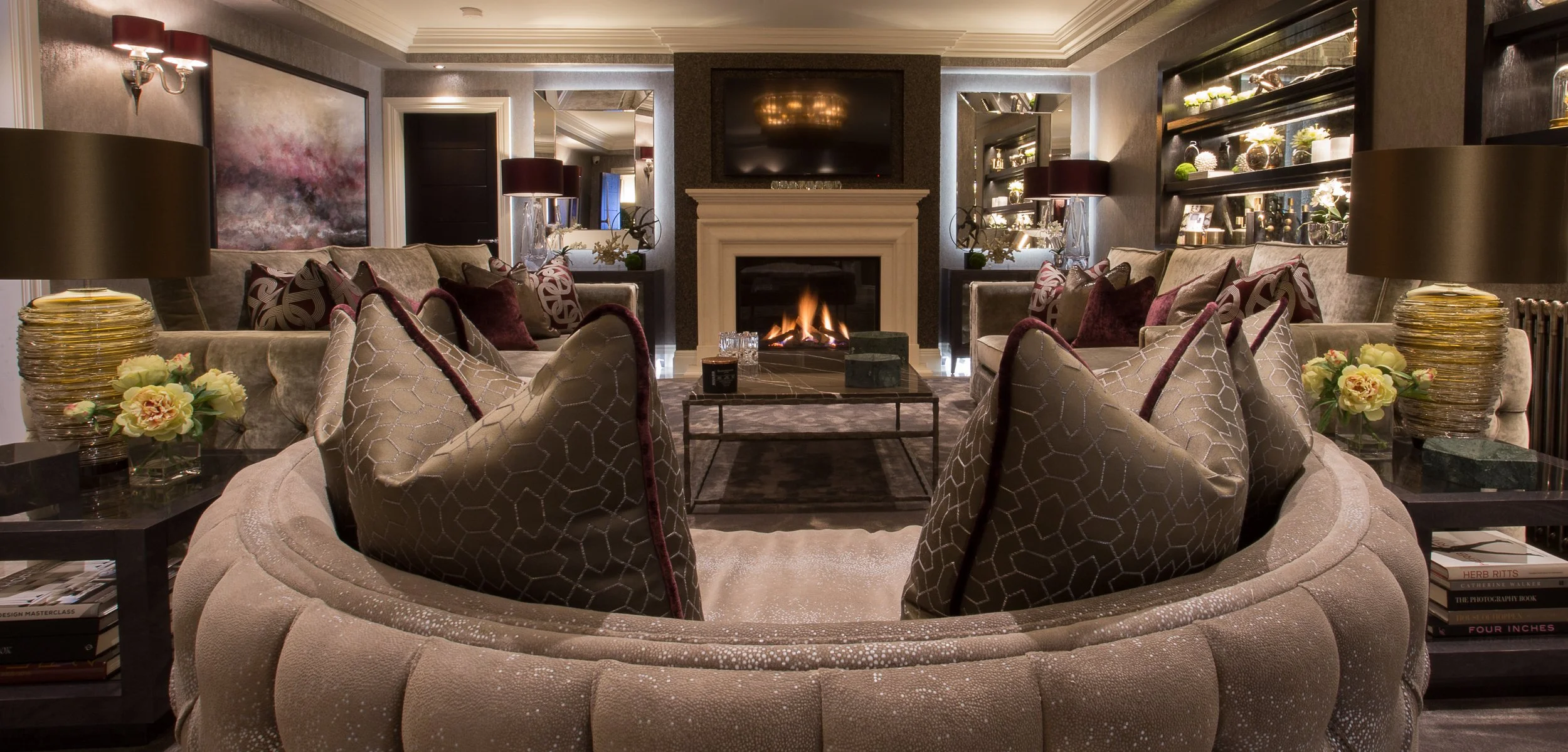 Elegant living room with a central fireplace, large TV above it, beige and burgundy color scheme, plush seating with patterned pillows, side tables with flowers, and built-in shelves with decorative items.