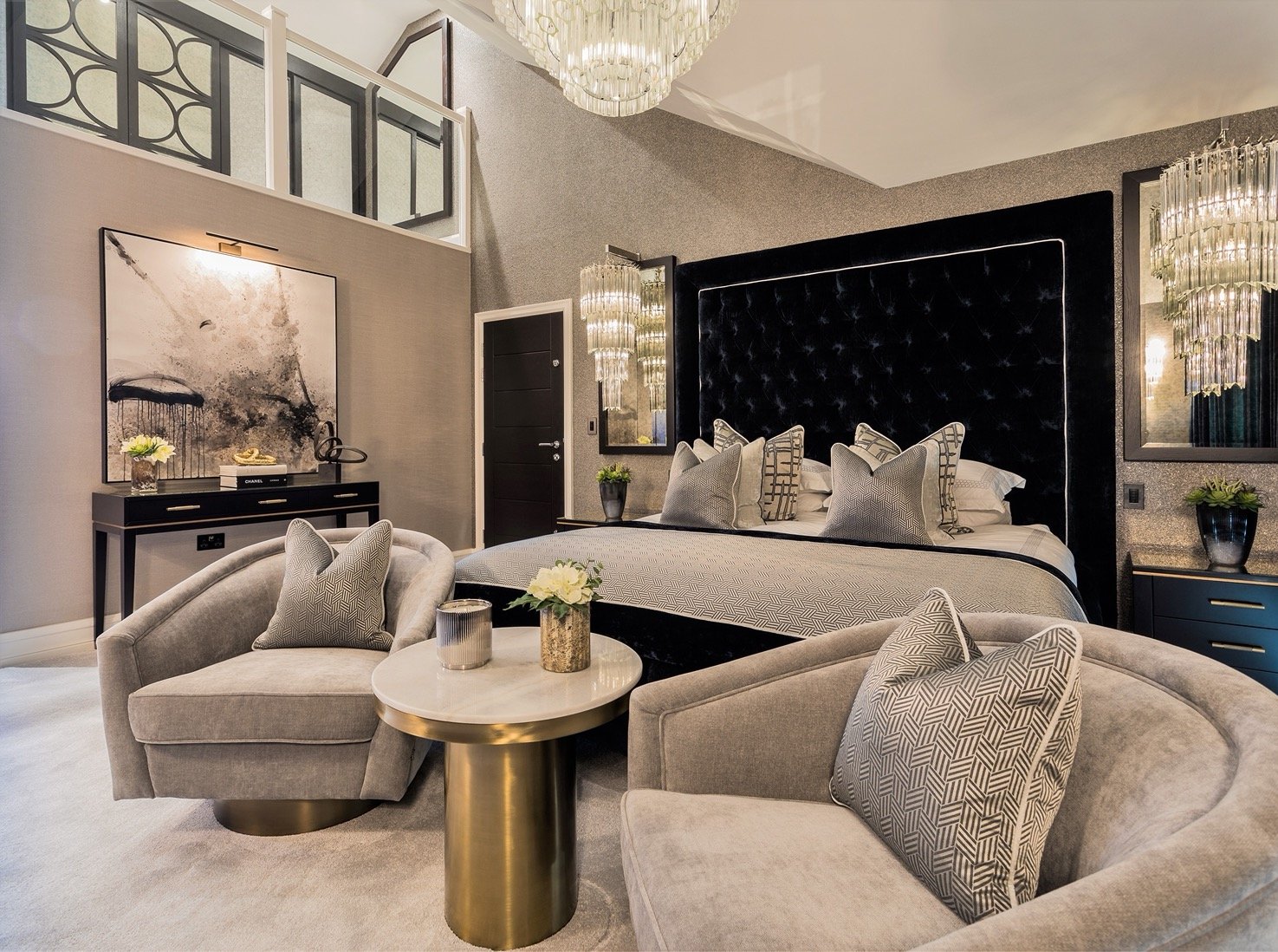 A luxurious bedroom with a large black tufted headboard, gray and patterned throw pillows, and matching bedding. Two plush light gray armchairs with decorative pillows face a round gold and white coffee table, which holds a small flower arrangement a