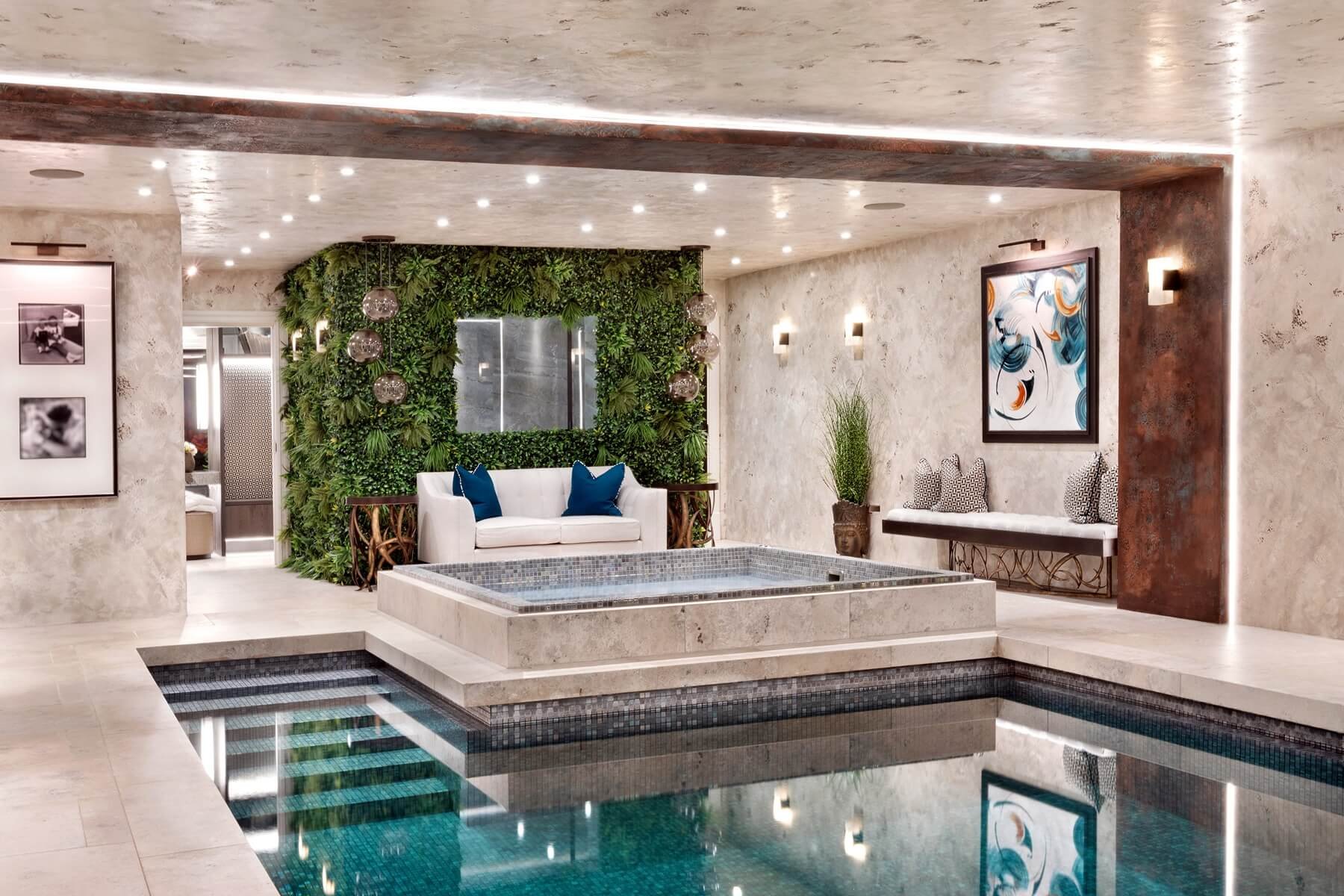 Indoor swimming pool area with beige stone walls, modern lighting, a lush green wall with plants, a white sofa with blue cushions, a bench with patterned pillows, framed abstract art, and a small potted plant.