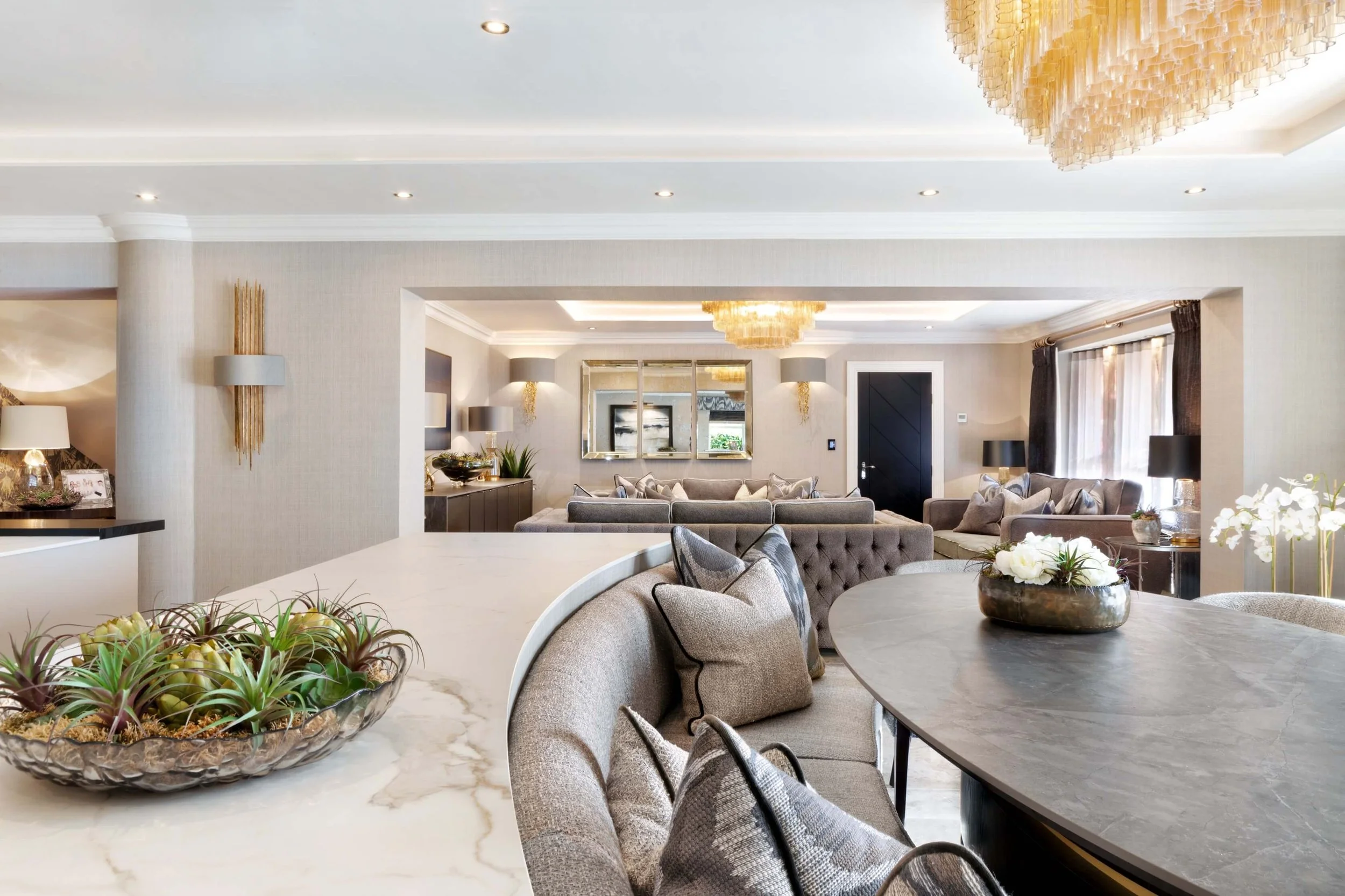 A spacious, modern living room with a large dining table, upholstered chairs, and a decorative centerpiece with plants. The background features a sitting area with sofas, lamps, and a mirrored wall, illuminated by chandeliers and natural light from w