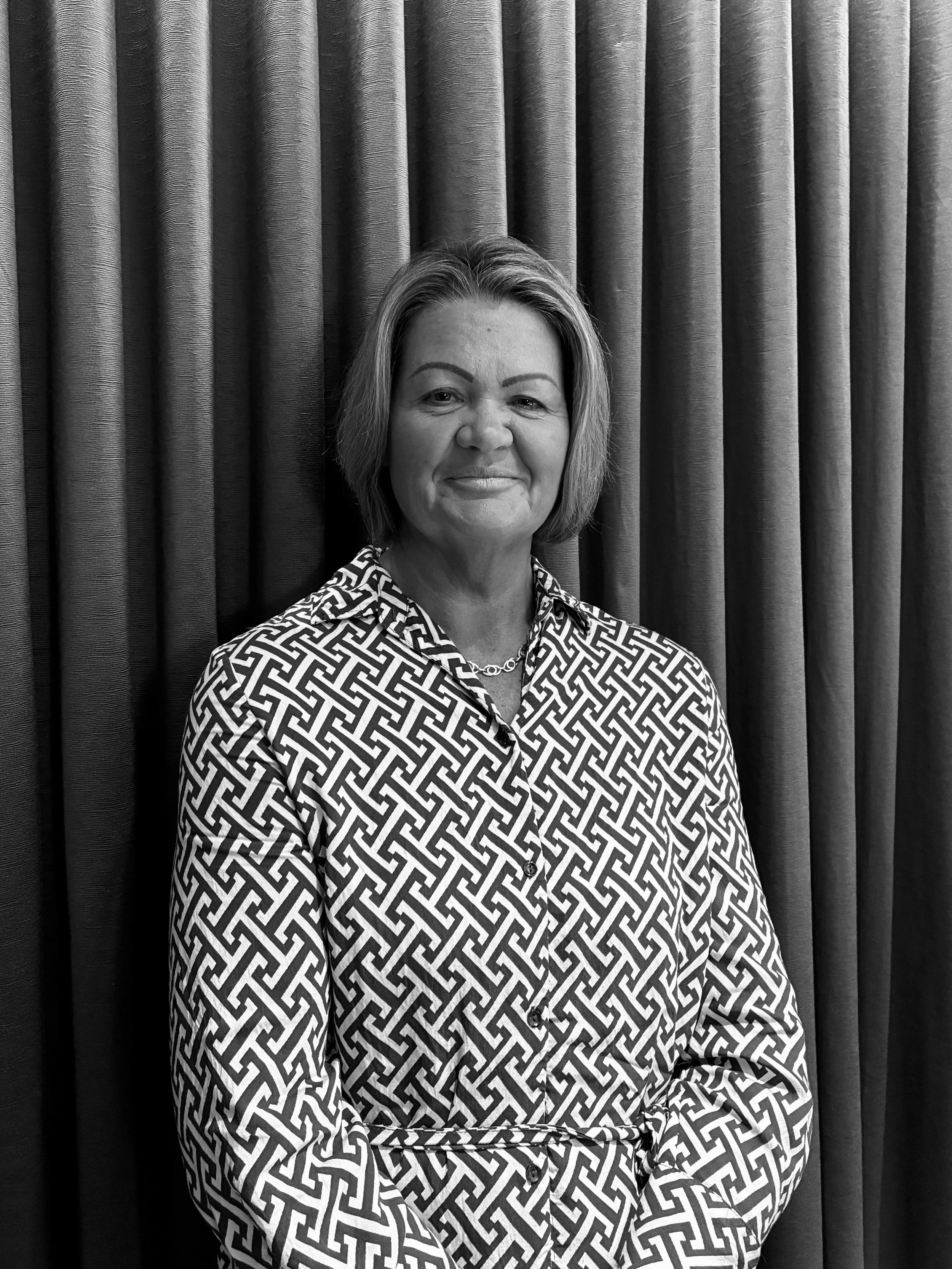 Black and white portrait of a woman with short hair, wearing a patterned shirt and a necklace, standing in front of a curtain with vertical folds.
