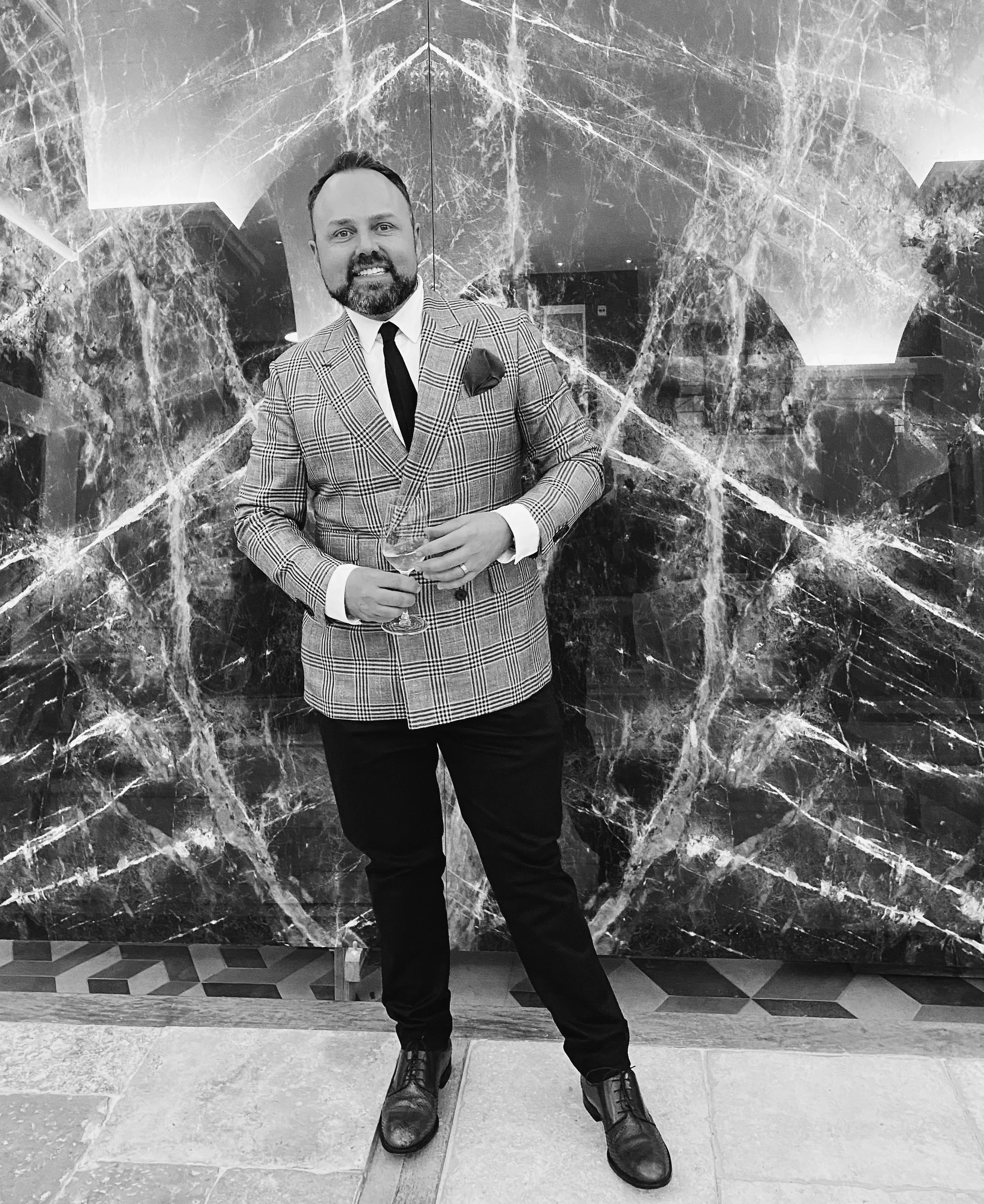 A man dressed in a plaid suit jacket, black pants, and black shoes, standing in front of a marble wall, holding a martini glass, smiling at the camera.