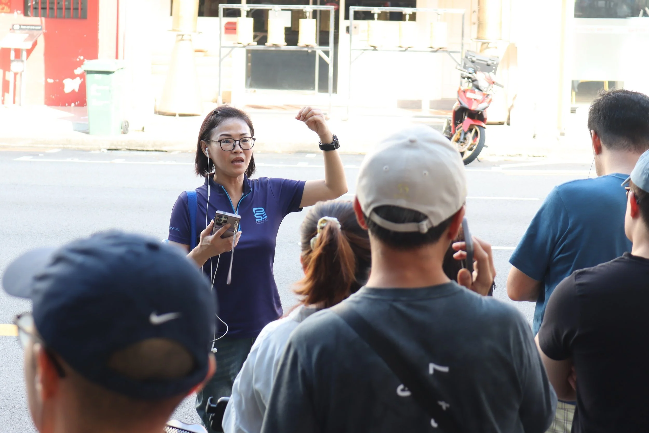 A woman in a blue shirt with a logo, wearing glasses and earphones, is speaking to a group of people outdoors on a street. She is holding a smartphone and gesturing with her right hand, possibly giving a presentation or leading a discussion.