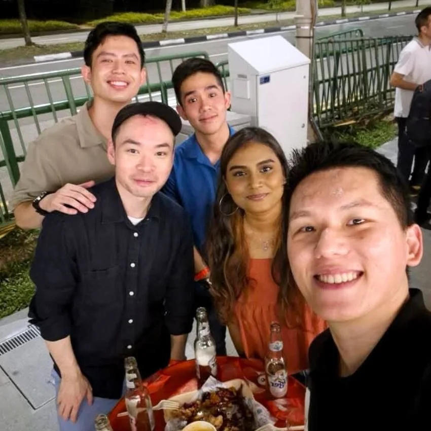 Group of five young adults taking a selfie outdoors at night, with a table of food and drinks in front of them, smiling at the camera.