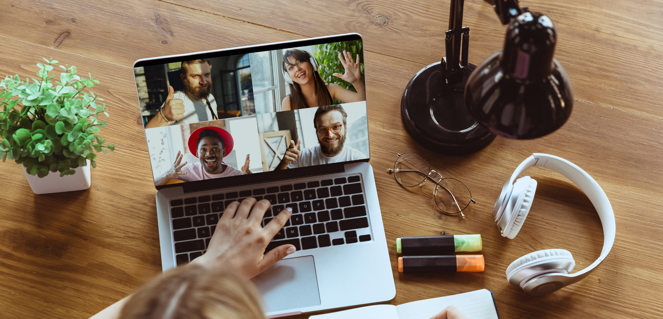 A person on a video call with four friends, with their hand hovering over a laptop keyboard. The laptop screen shows four participants, each smiling and one giving a thumbs up or wave. The workspace includes a potted plant, glasses, headphones, highlighters, a notebook, and a desk lamp on a wooden desk.