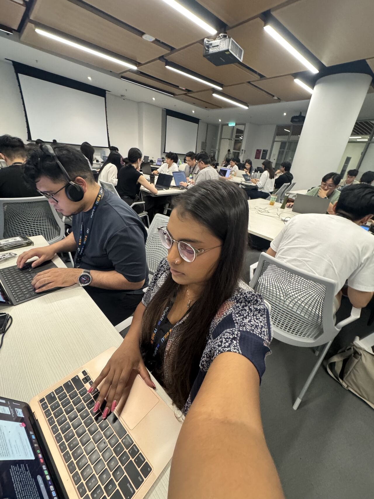 People working on laptops in a modern conference room with large displays, some wearing headphones, engaged in a professional event or workshop.