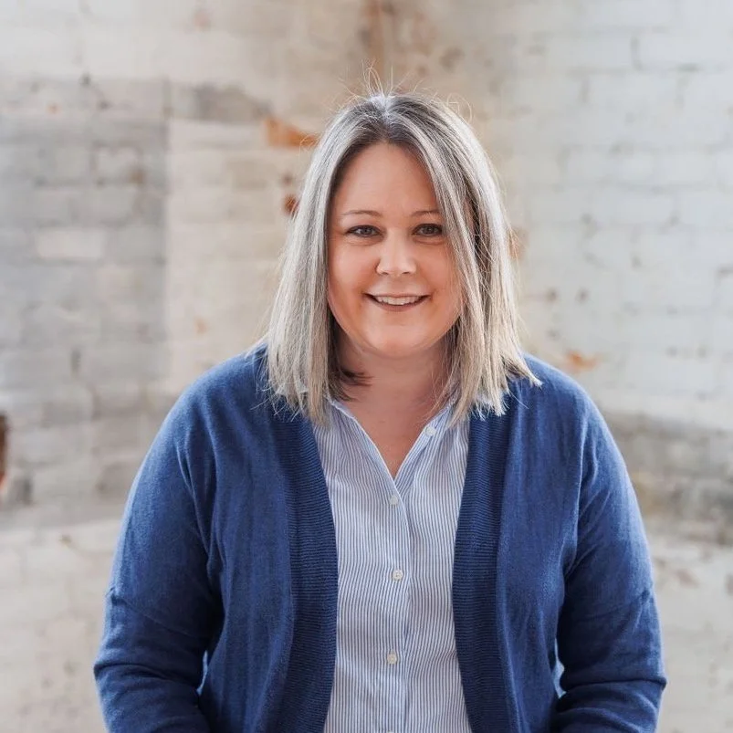 Person with shoulder-length hair wearing a blue cardigan and striped shirt against a light brick background.