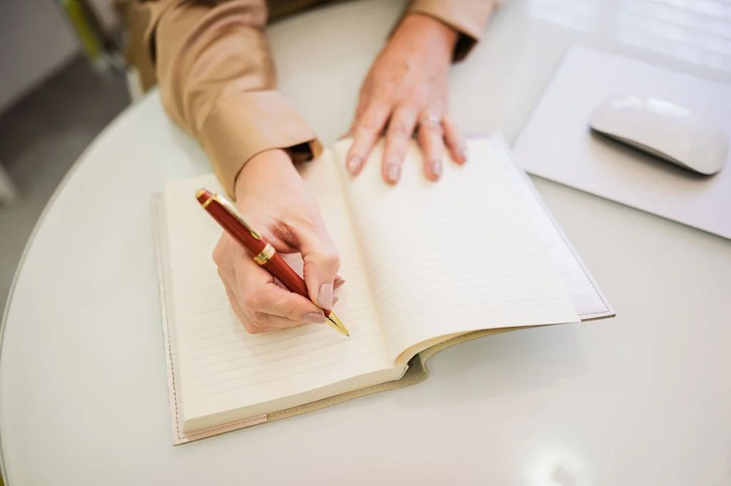 Person writing in a lined notebook with a red pen on a white desk, next to a computer mouse.